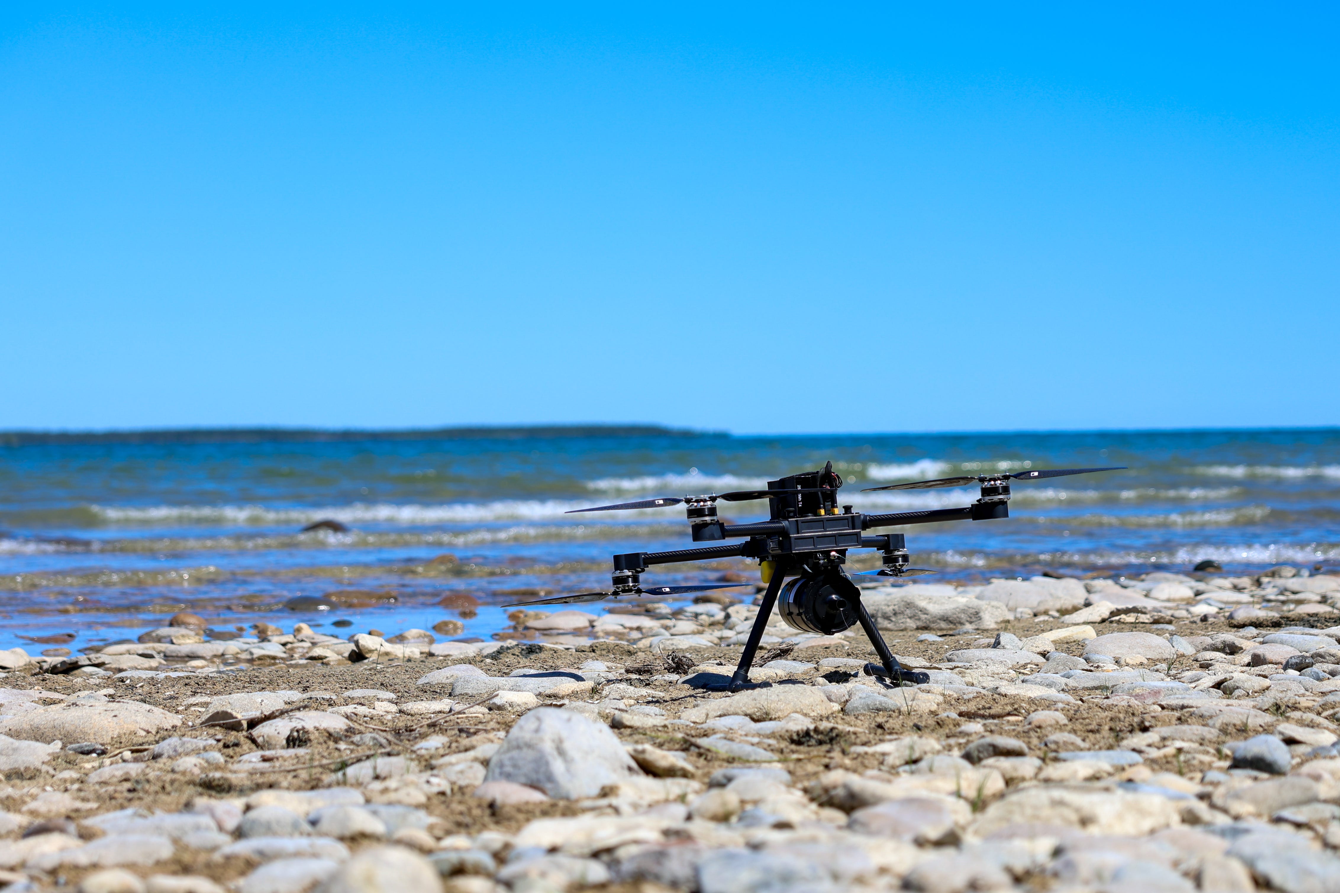 A small, black fiber-optic first-person view drone sits on rocks at a beach, with waves and a blue sky in the background.