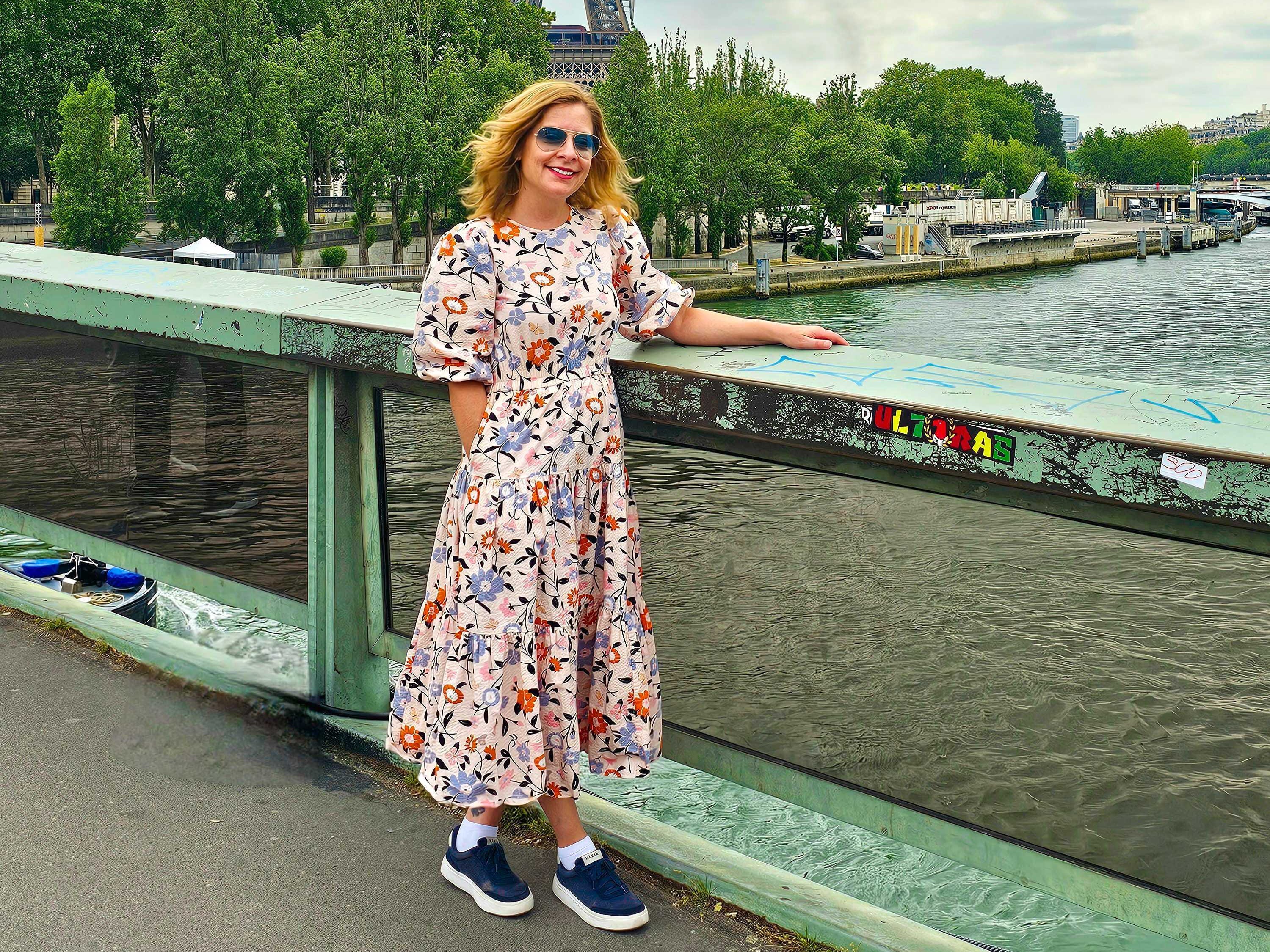 The writer wears a blue and red floral dress and walks along a bridge with a green railing in front of the Eiffel Tower