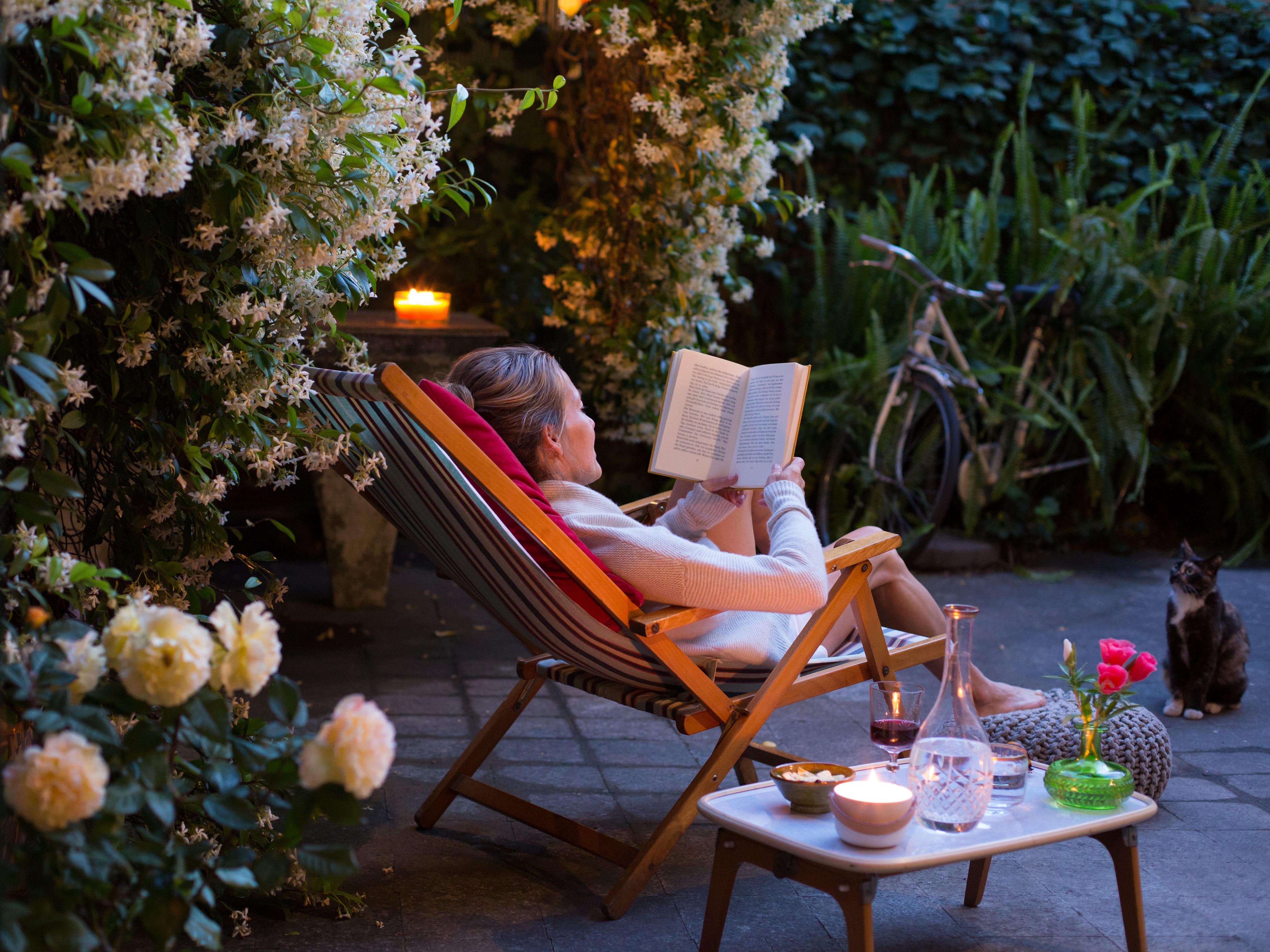 A woman reading a book in a backyard with a glass of wine and snacks.