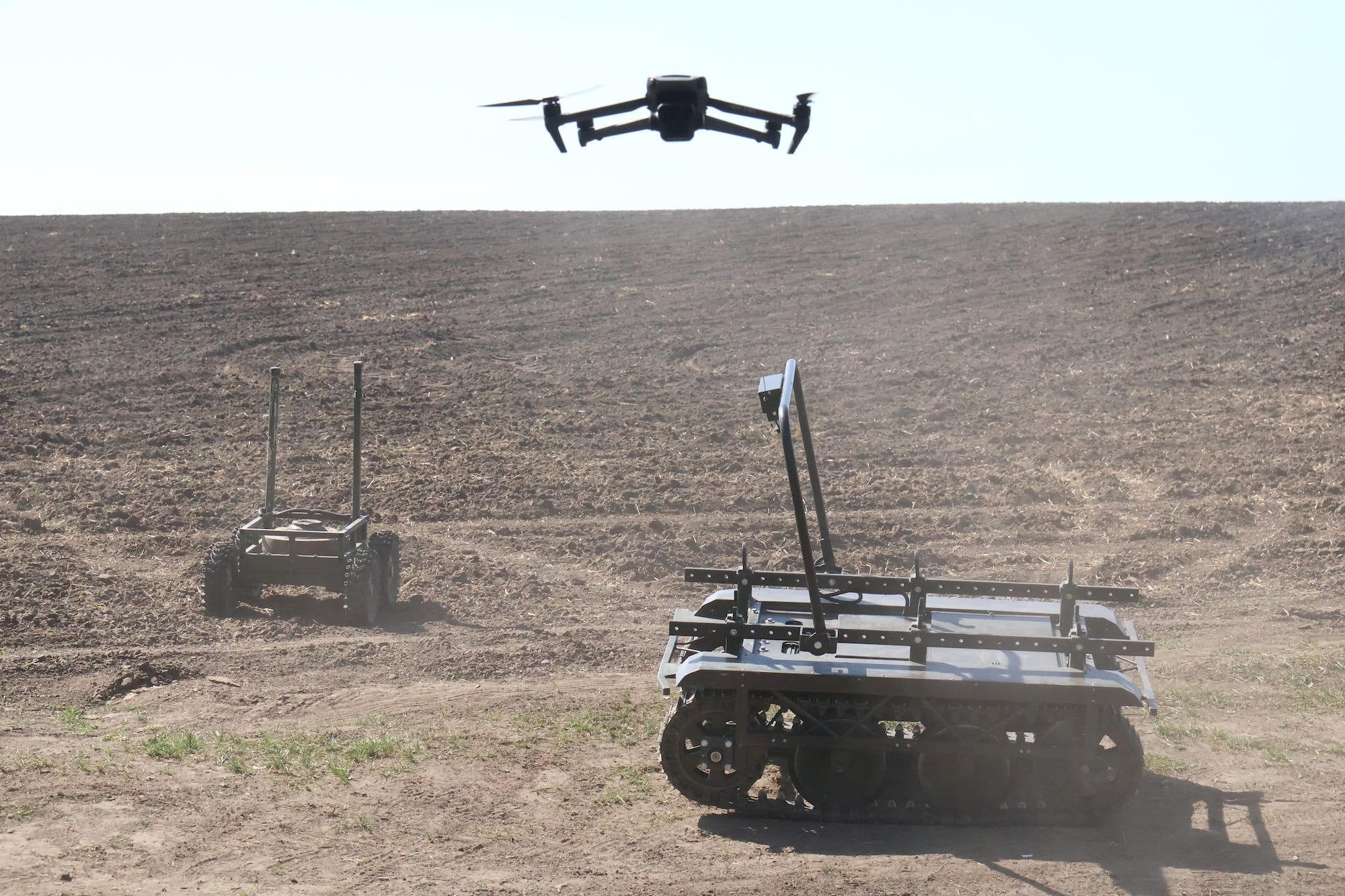 Two ground robots with tracks on brown ground with a drone in the sky above them