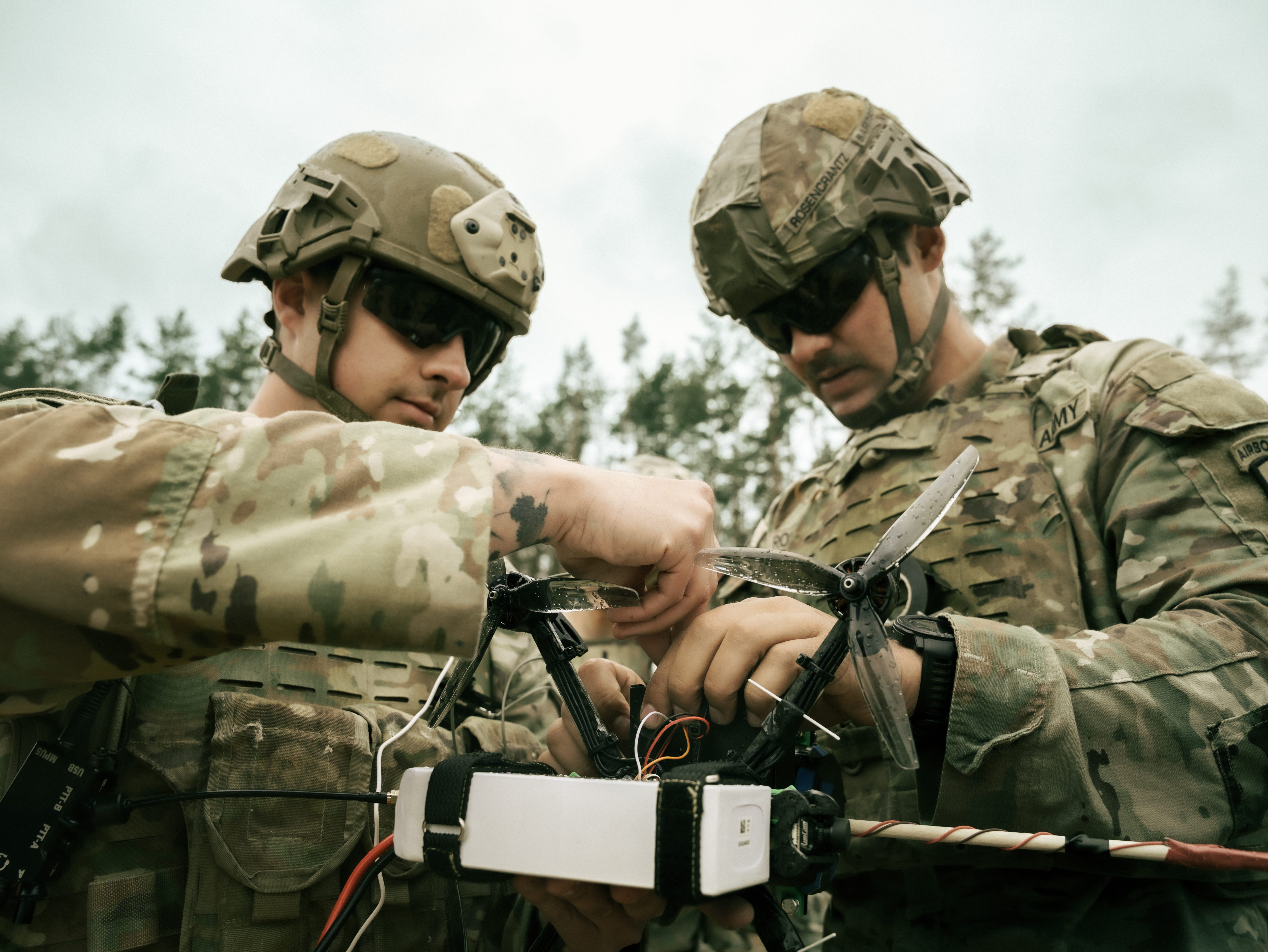 Two US Army soldiers wearing camouflage and sunglasses work on a small drone. The sky is overcast in the background.