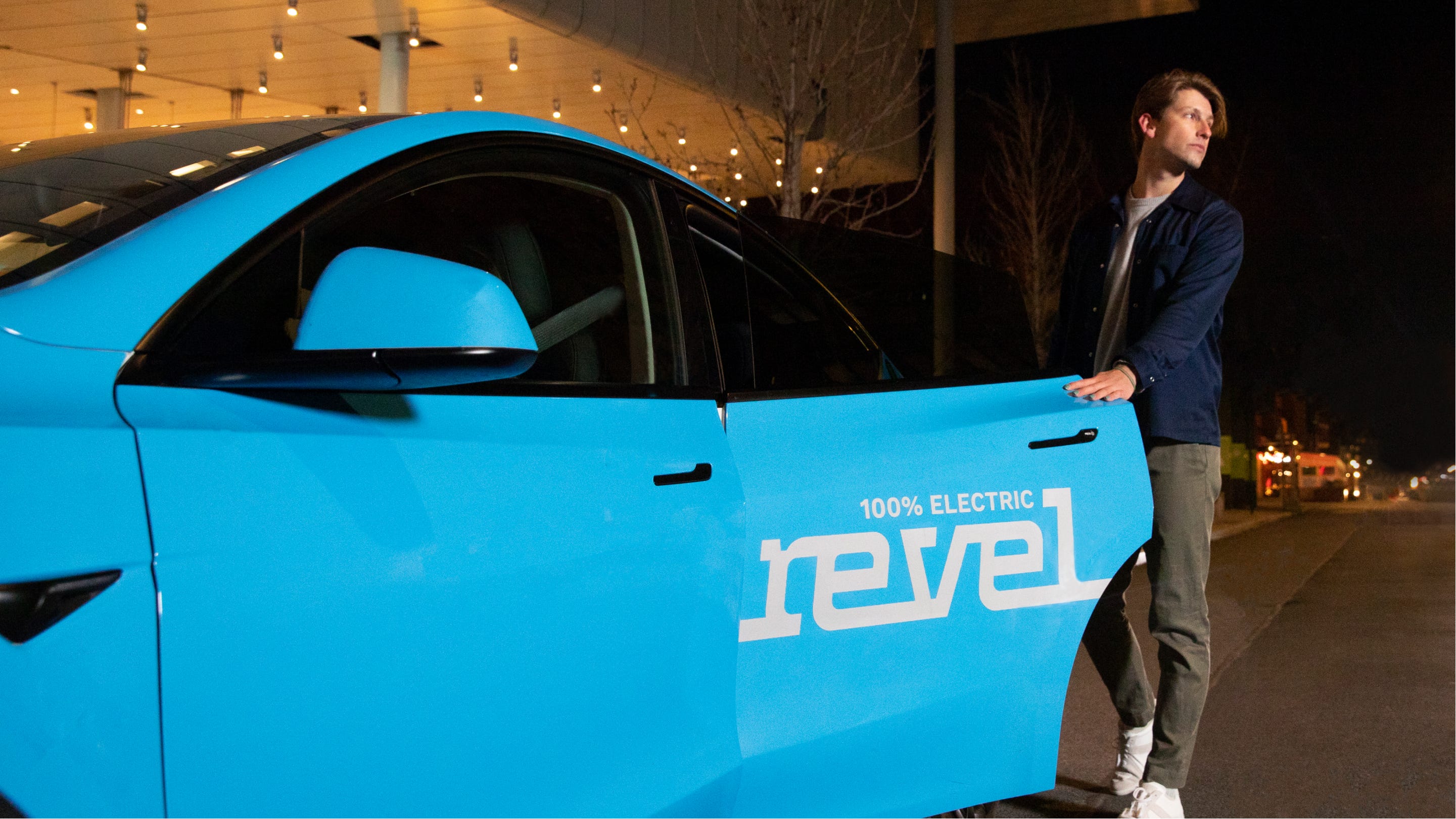 A man wearing a blue jacket and green pants opens the door to a Tesla car owned by Revel. The car is painted light blue and the writing on the door says that it's a 