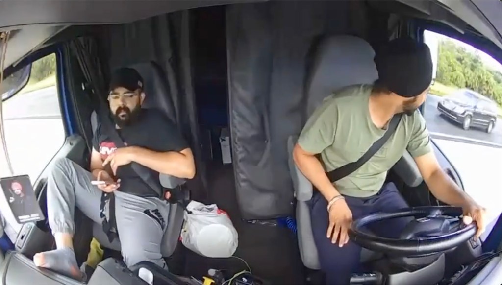 Interior view of a semi-truck cab with two men, one driving and one on a phone.