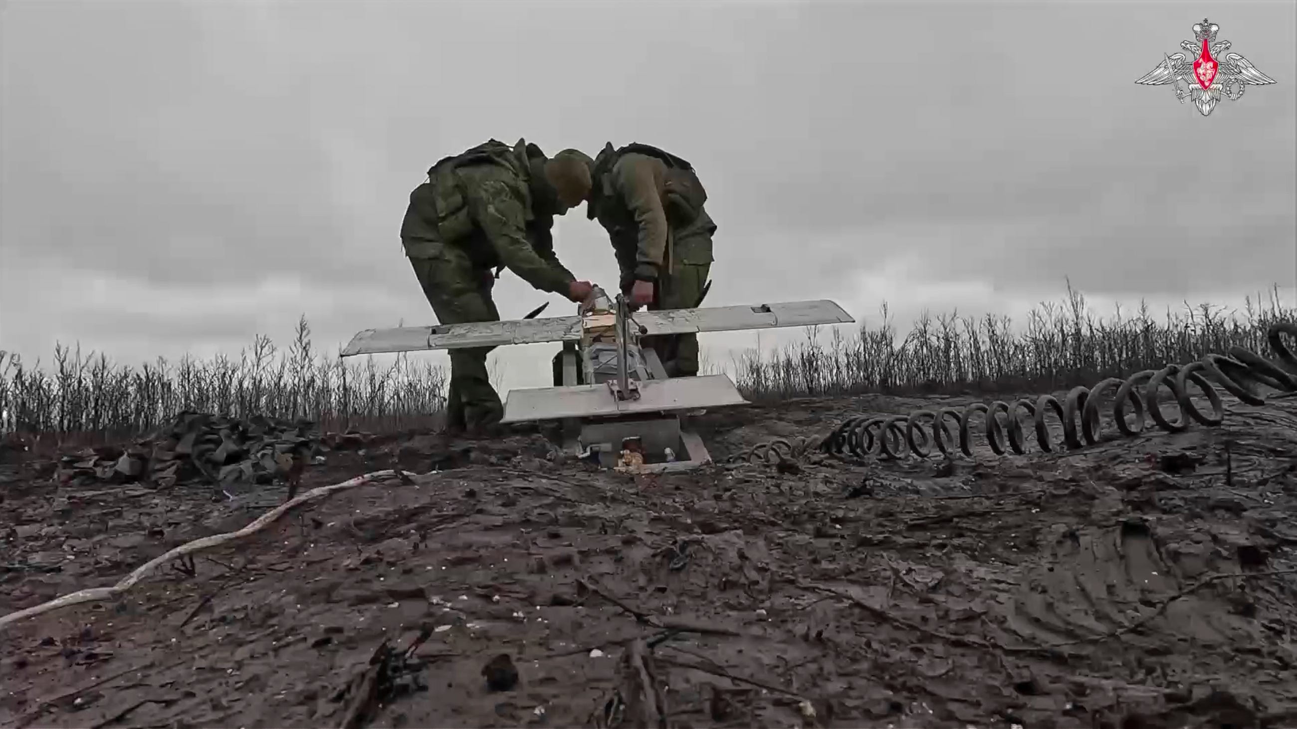 Two men hold a large grey drone against a grey sky and a muddy ground