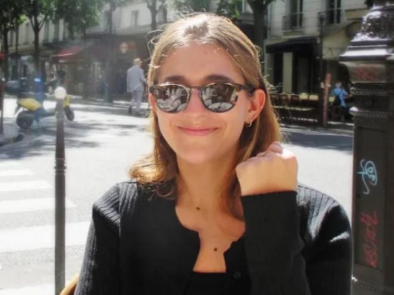 A woman sitting outside, smiling and wearing sunglasses.