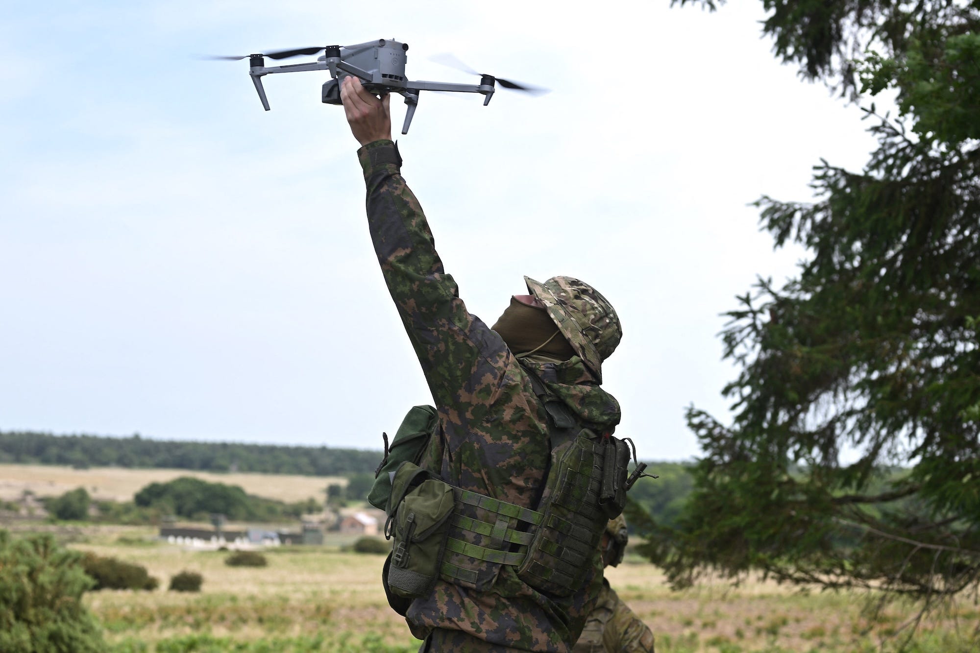 A man in camouflage clothing and a hat and with a green balaclava covering his face holds a grey drone in the air with grass and trees behind him