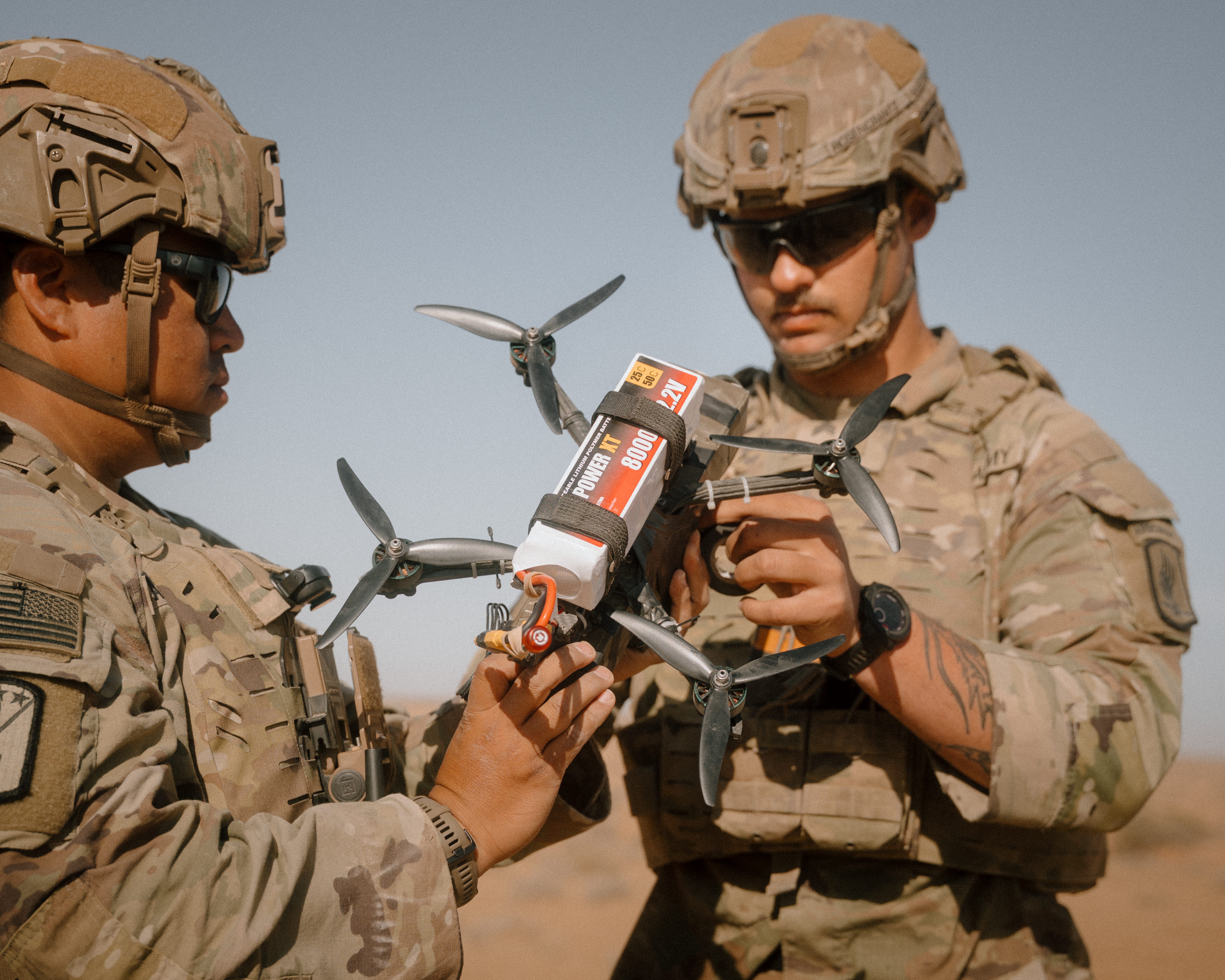 Two US Army soldiers wearing camouflage are holding and examining a small quadcopter drone. The sky is grayish blue in the background.
