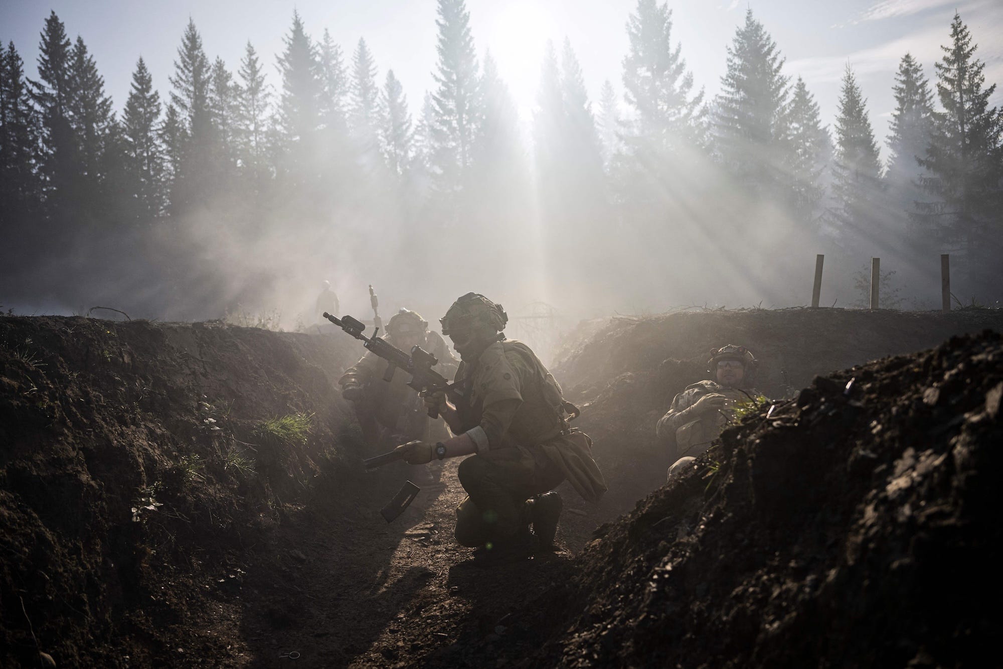 A man holding a firearm in a trench with mist and trees behind him