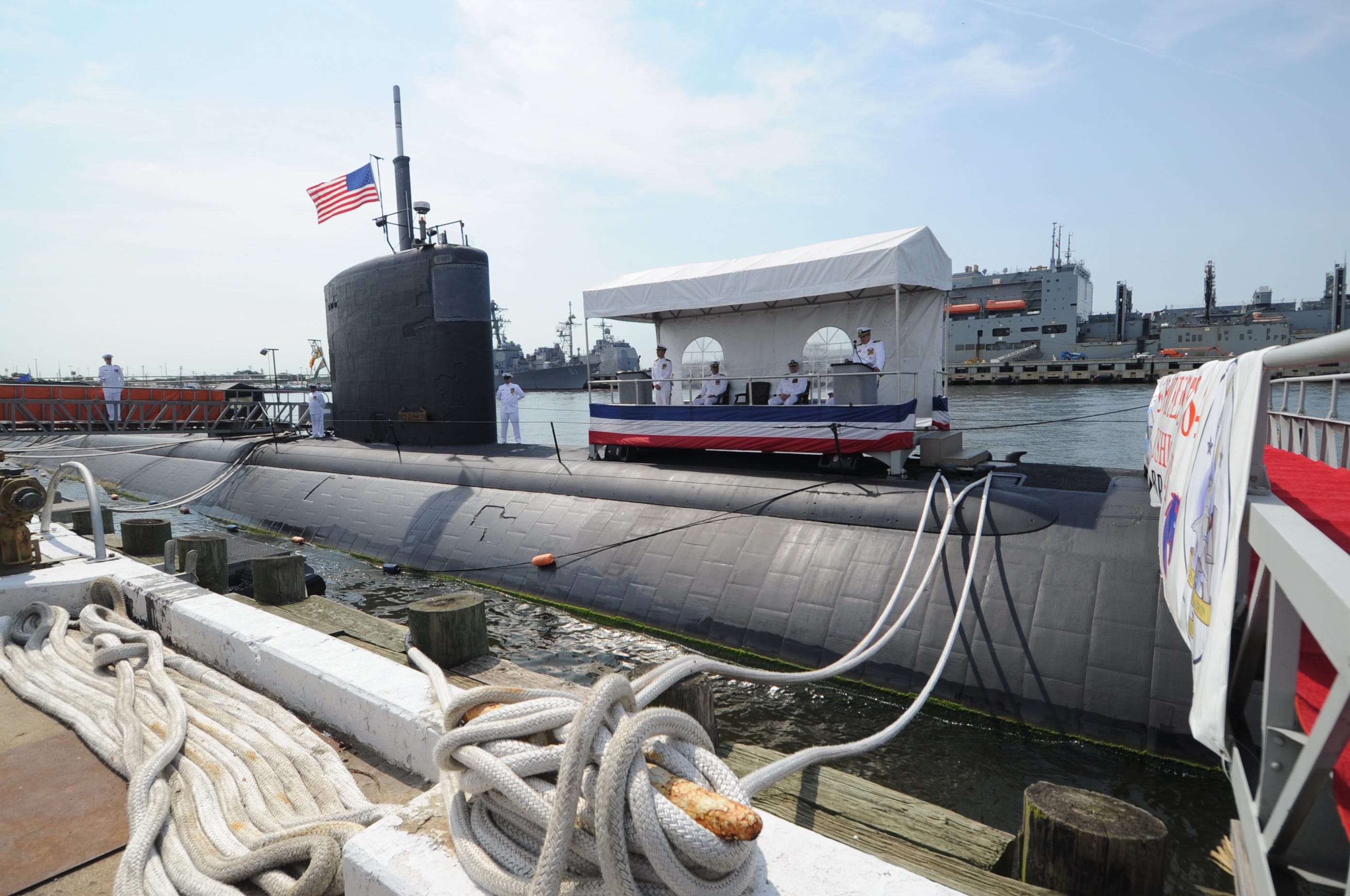 The USS Boise submarine sits in the water with sailors standing around and on top of it. On top of the submarine, a stage sits with some Navy officials sitting and standing on it. The sky is blue and cloudy in the background.
