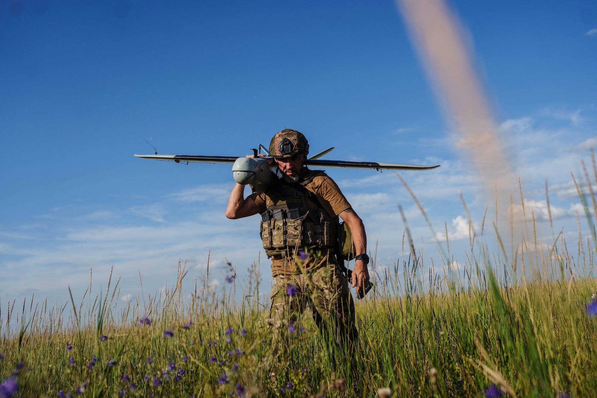 A man stands in a green field under a blue sky wearing green camouflage gear and holding a drone on his shoulder