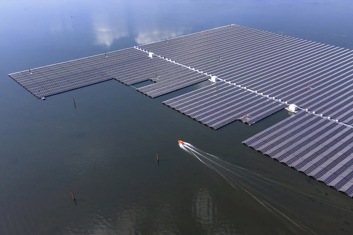 An aerial view shows a sprawling expanse of solar panels as they float on open water. A small a motor boat passes by the field of panels, leaving a wake behind it.