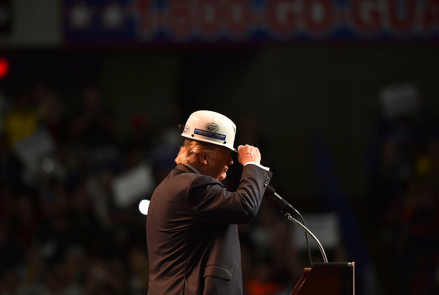 Donald Trump is seen in profile as he tips a coal miners hard-hat toward a crowd, out of view, while standing in front of a microphone.