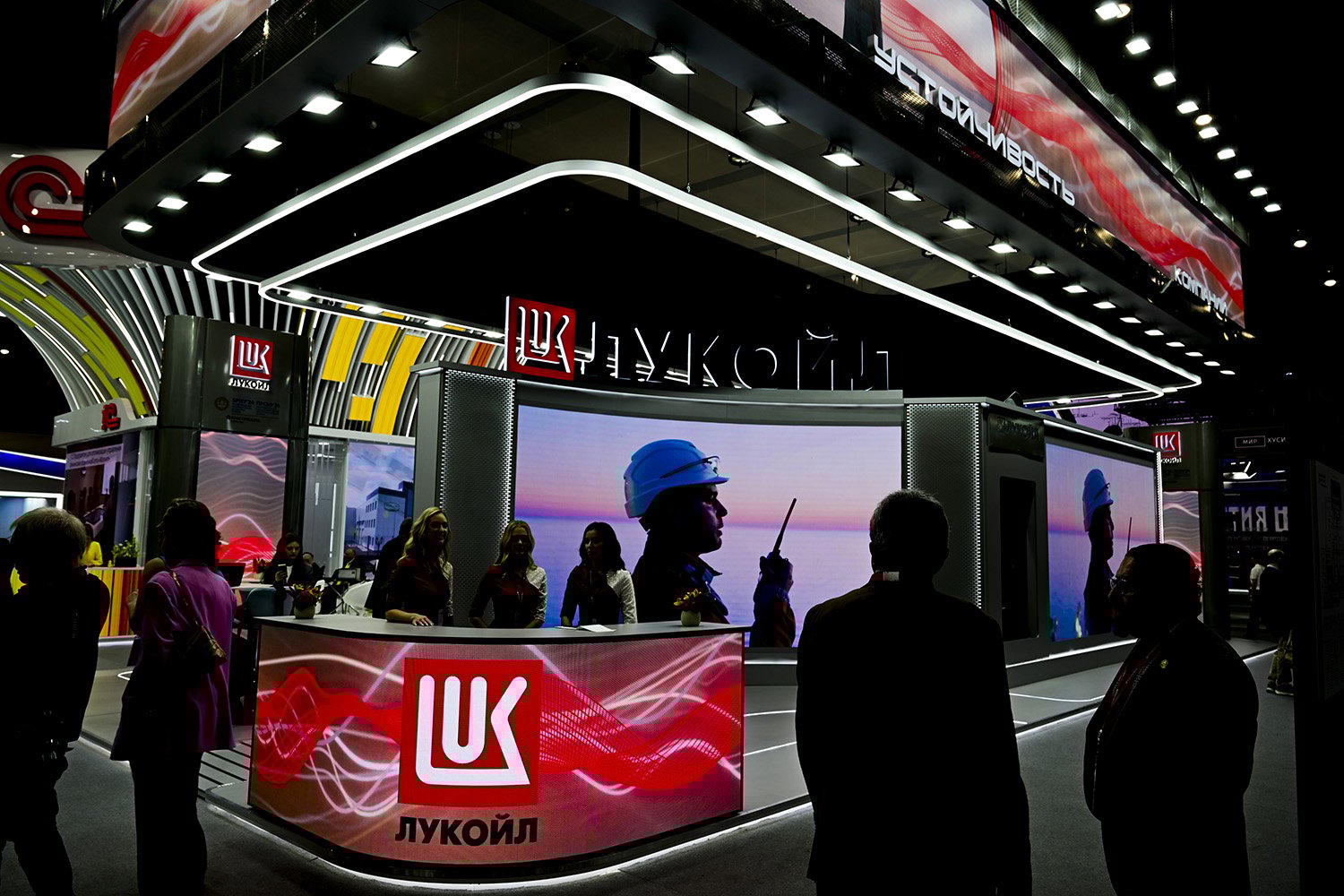 A screen shows a worker in a hard hat behind the table at a Lukoil booth. People are silhouetted as they walk along a concourse in front of the booth; three women sit at the table, which is fronted with another screen, this one showing Lukoil's logo in red.