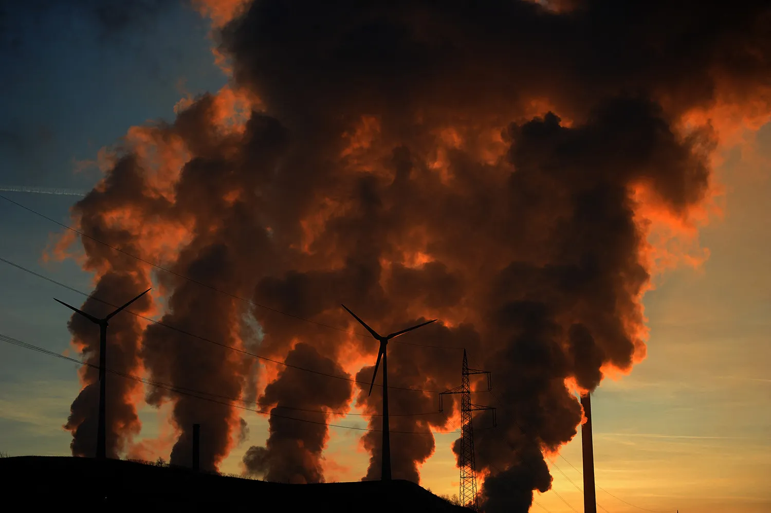 Orange-black columns of steam or smoke rise from cooling towers. Wind turbines are silhouetted against the plumes rising behind them. Beyond all this is a blue sky lit up with orange splotches, likely at either dawn or dusk.