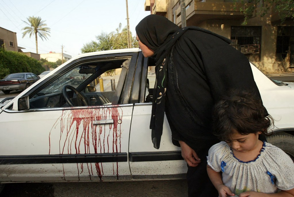 Blood stains on a white car; a woman and child stand nearby.