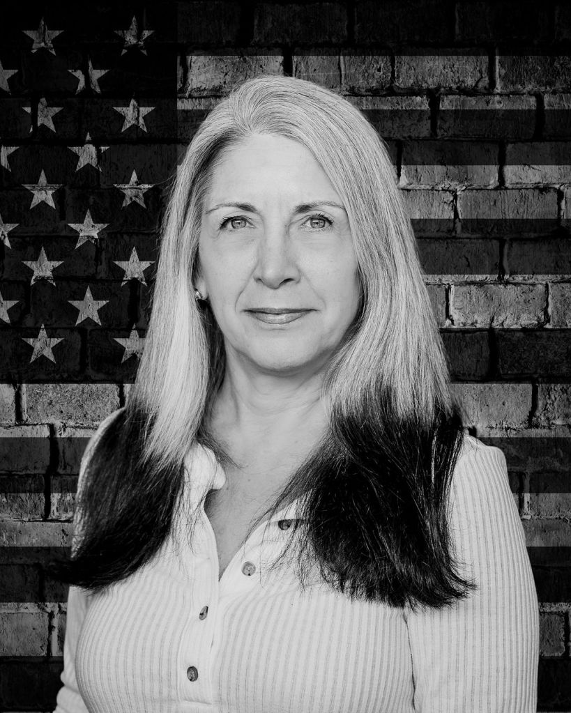 Black and white photo of Gina Keating in front of a brick wall depicting the American flag.