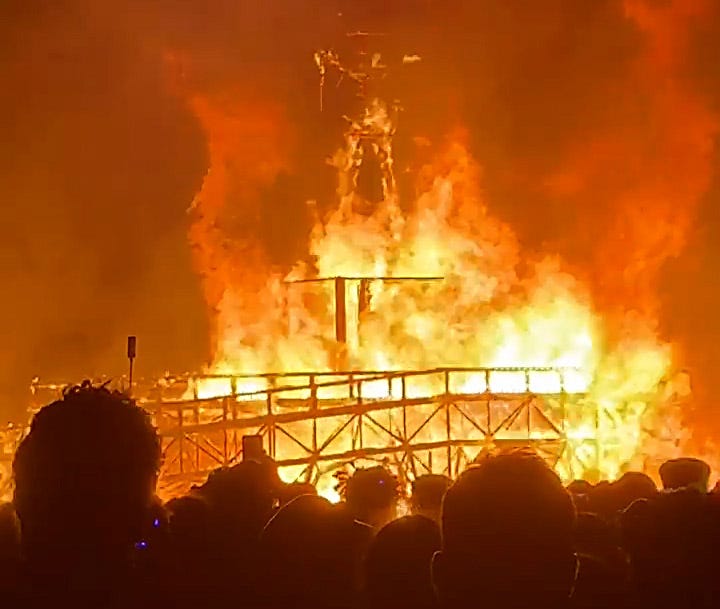 Large structure engulfed in flames, viewed from a crowd.