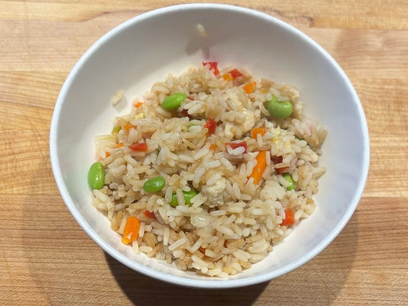 A bowl of Walmart's Great Value chicken fried rice with visible carrots, edamame, and red pepper in a white bowl on a wooden cutting board.