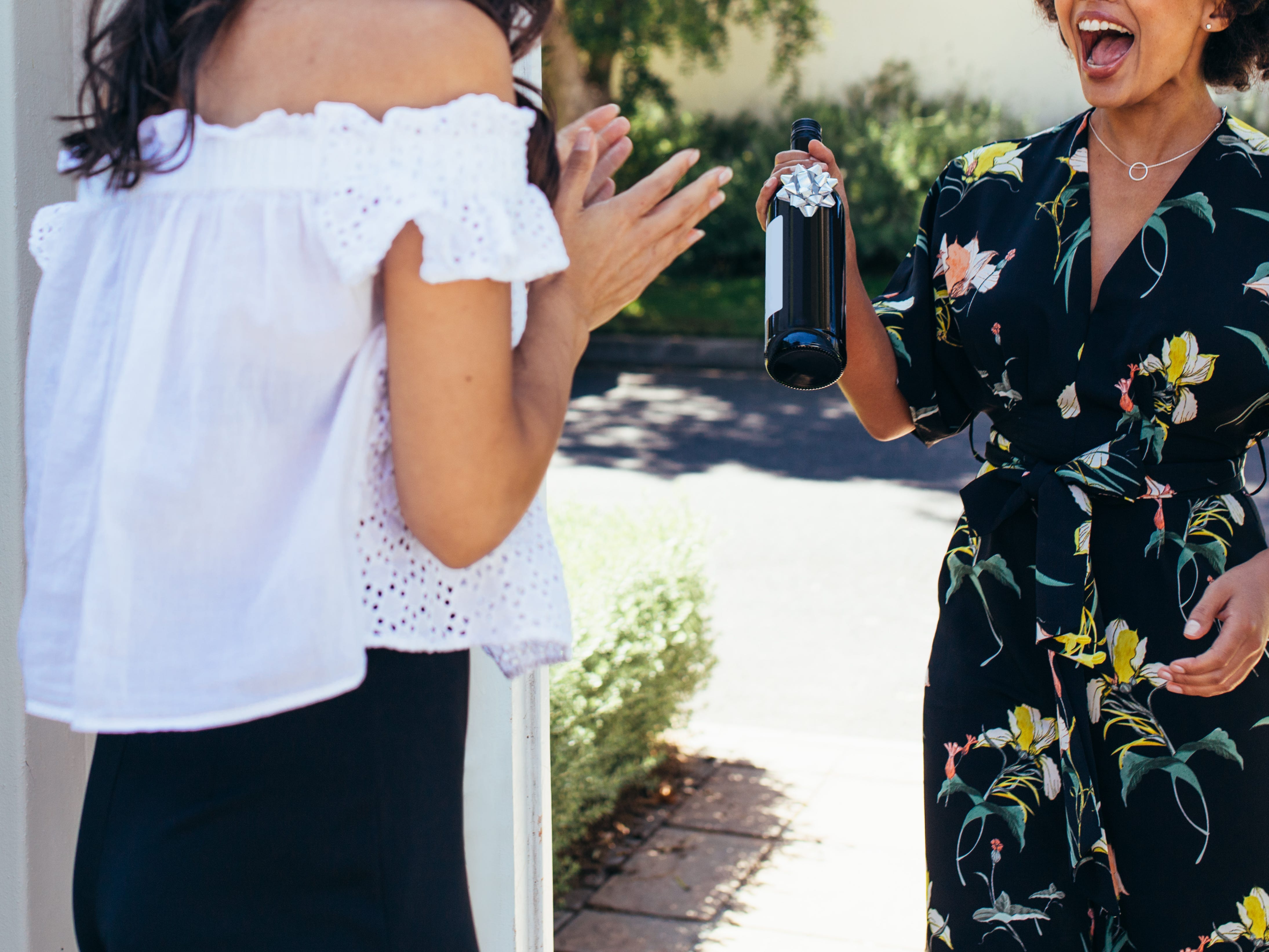 A woman welcoming a guest into the doorway of her home, as the guest carries a bottle of wine with a bow on it.