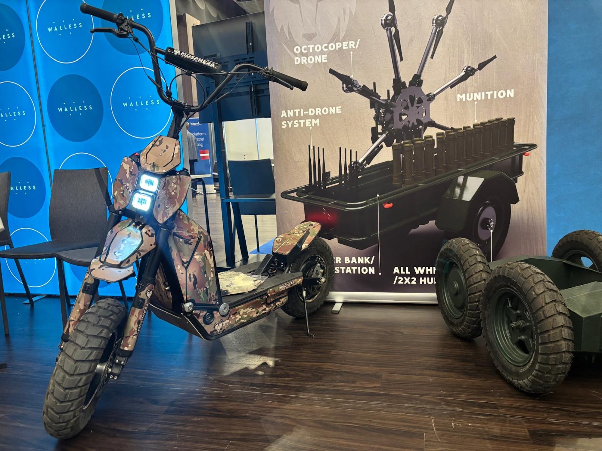 A camouflage-colored scooter with large wheels on a wooden floor in front of a promotional banner for another military system