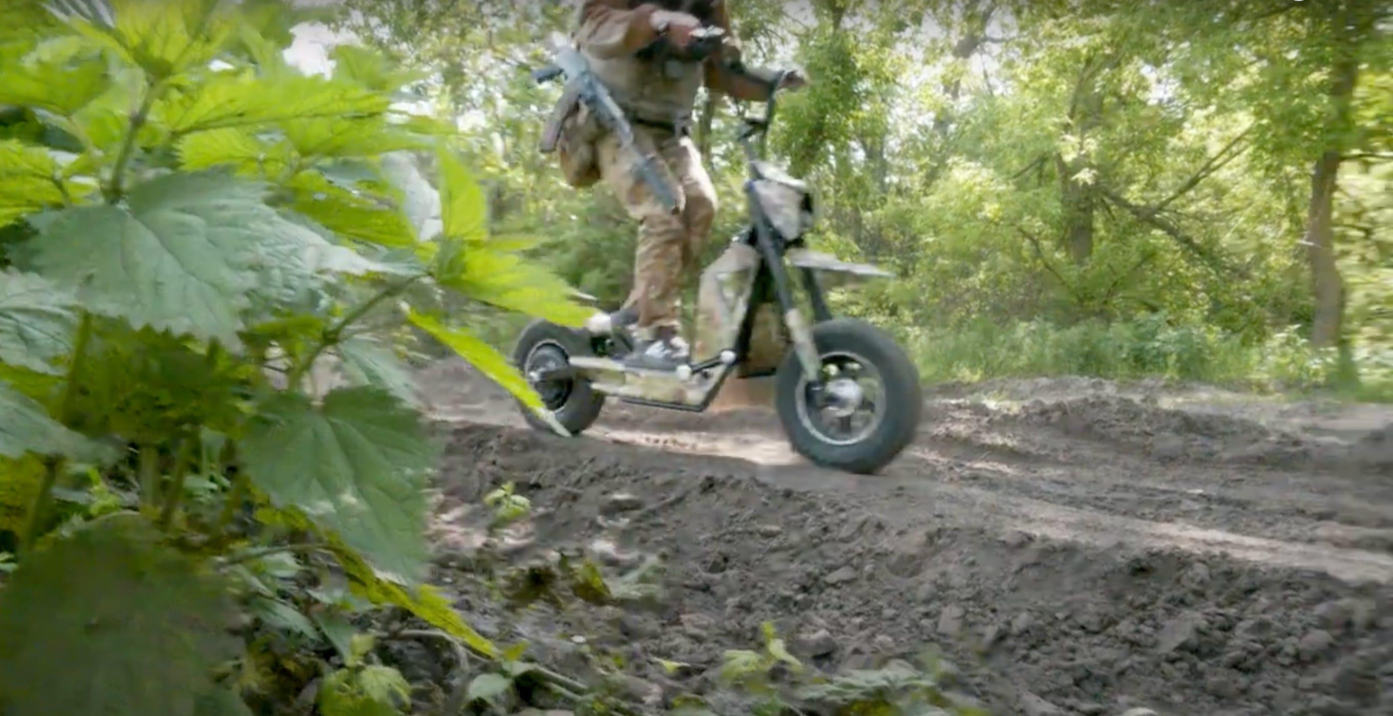 A man in camouflage gear on a scooter drives on a dirt road with green foliage on either side