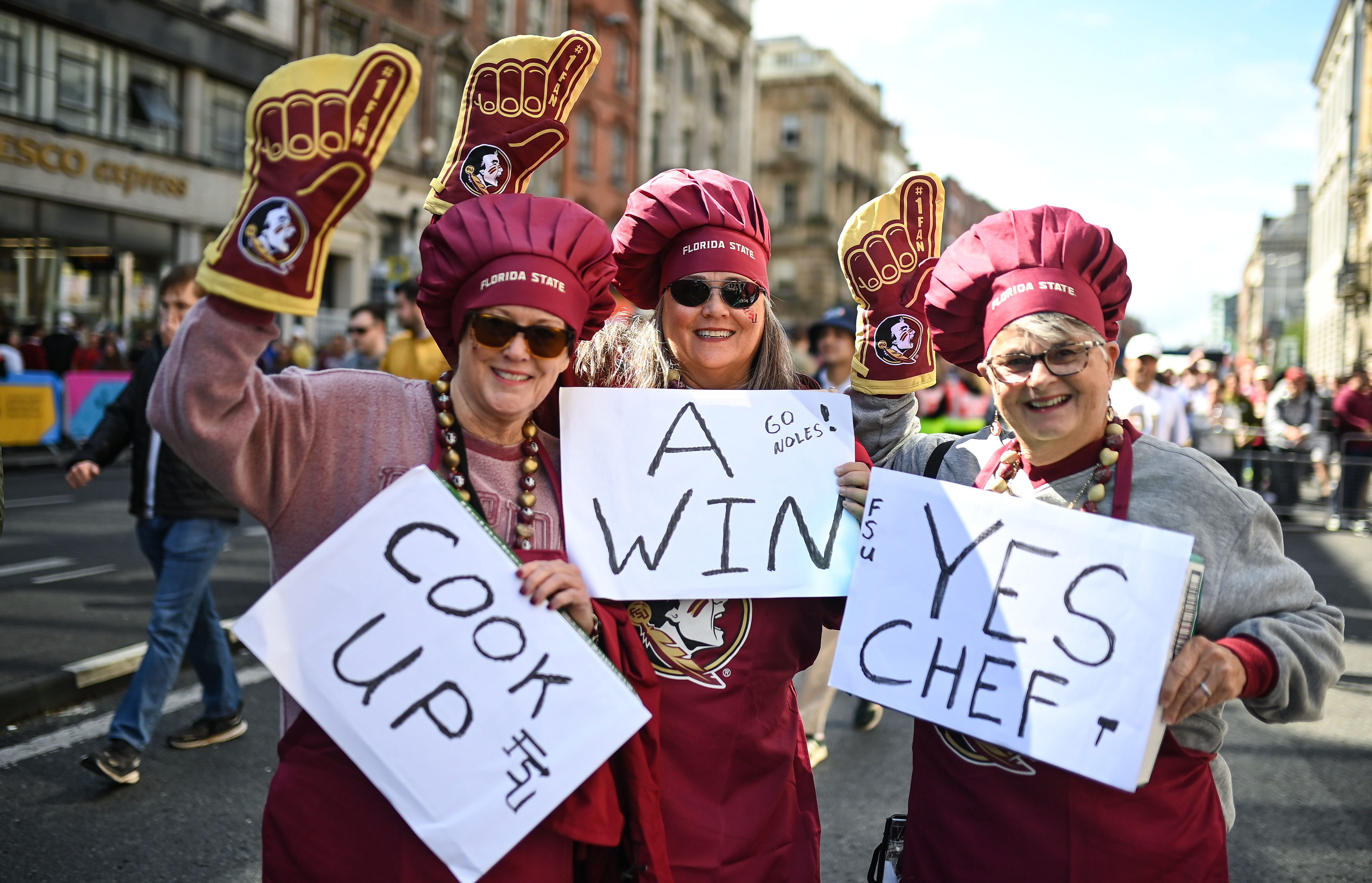 Three women wearing Florida State merch, including chef hats and aprons, holding signs that say "cook up," "a win," and "yes chef."