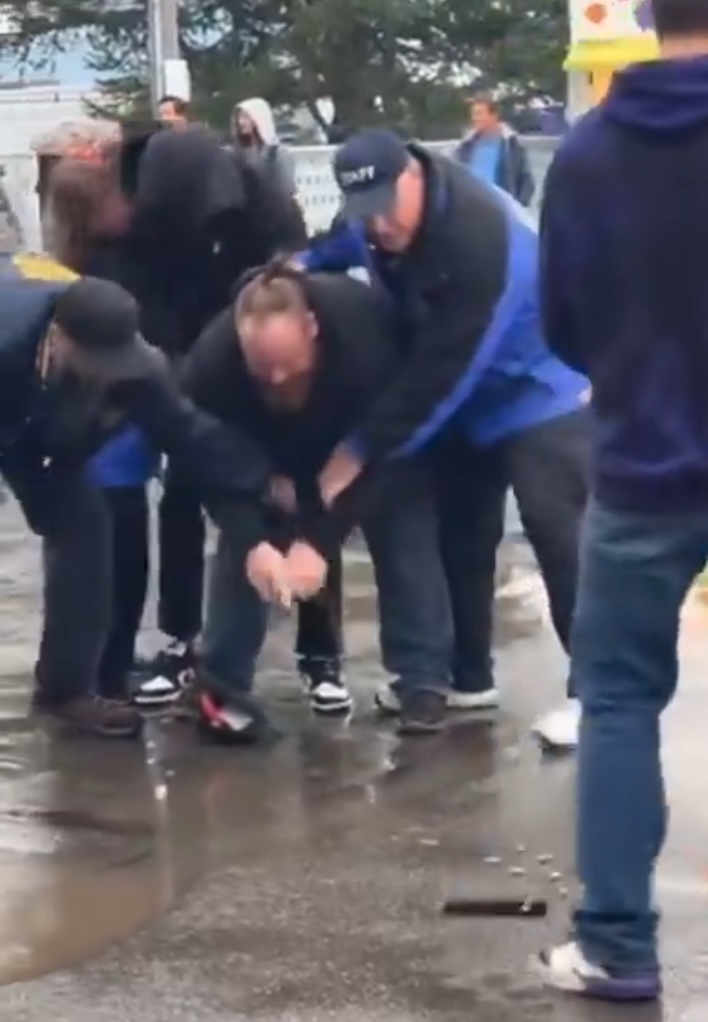 A man being held down by several people while he appears to be picking up something from the ground.