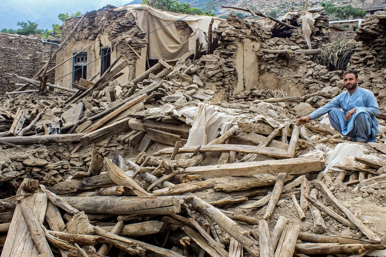 An Afghan man wearing blue sits on top of the remains of a house following an earthquake.