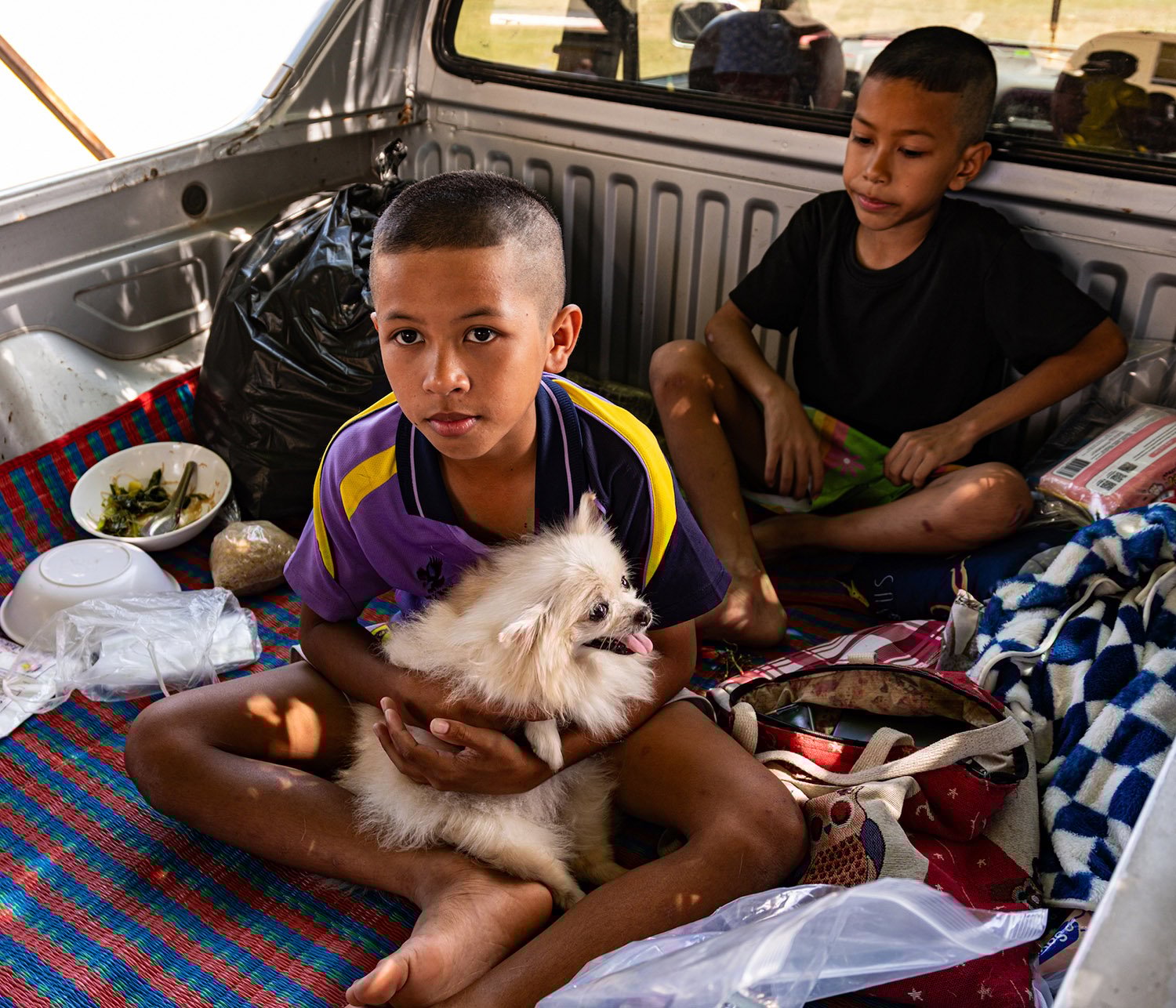 Two boys sit on blankets next to bowls of food in the back of a pickup. One holds a small fluffy dog.