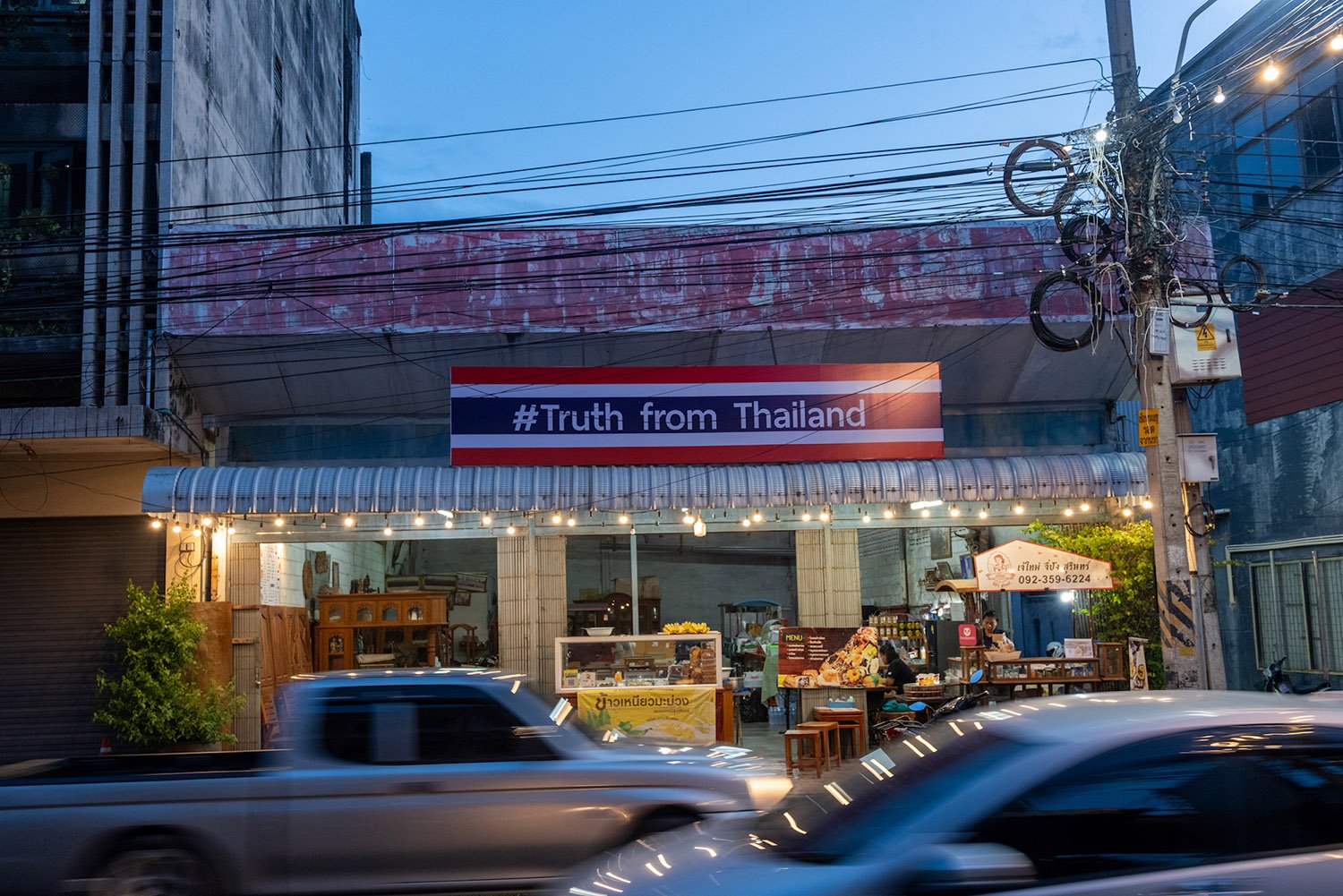 A banner that reads #Truth from Thailand hangs on a storefront with powerlines above and cars passing in front.