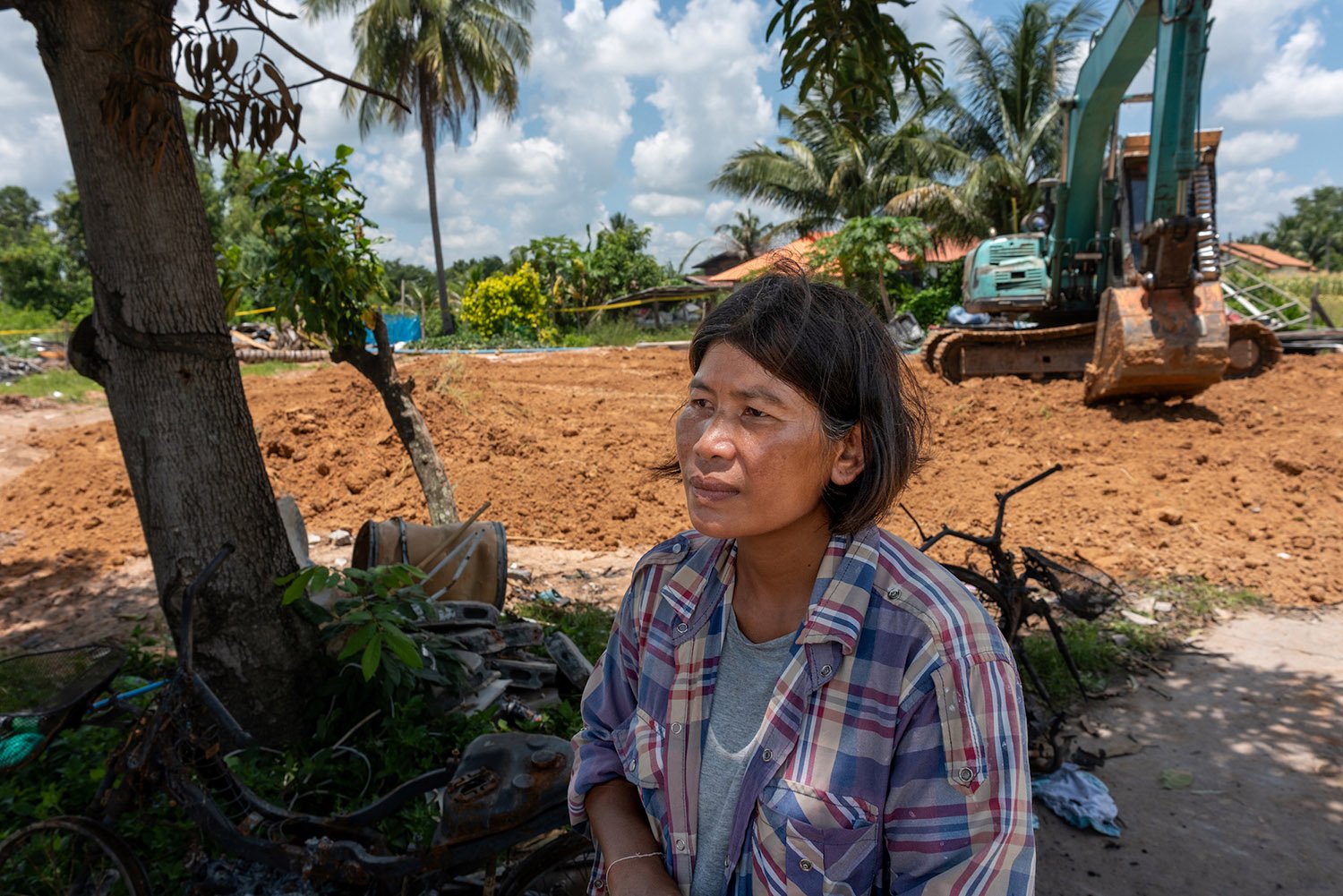 A person in a plaid shirt stands in front of a dirt covered lot with an excavator and palm trees.