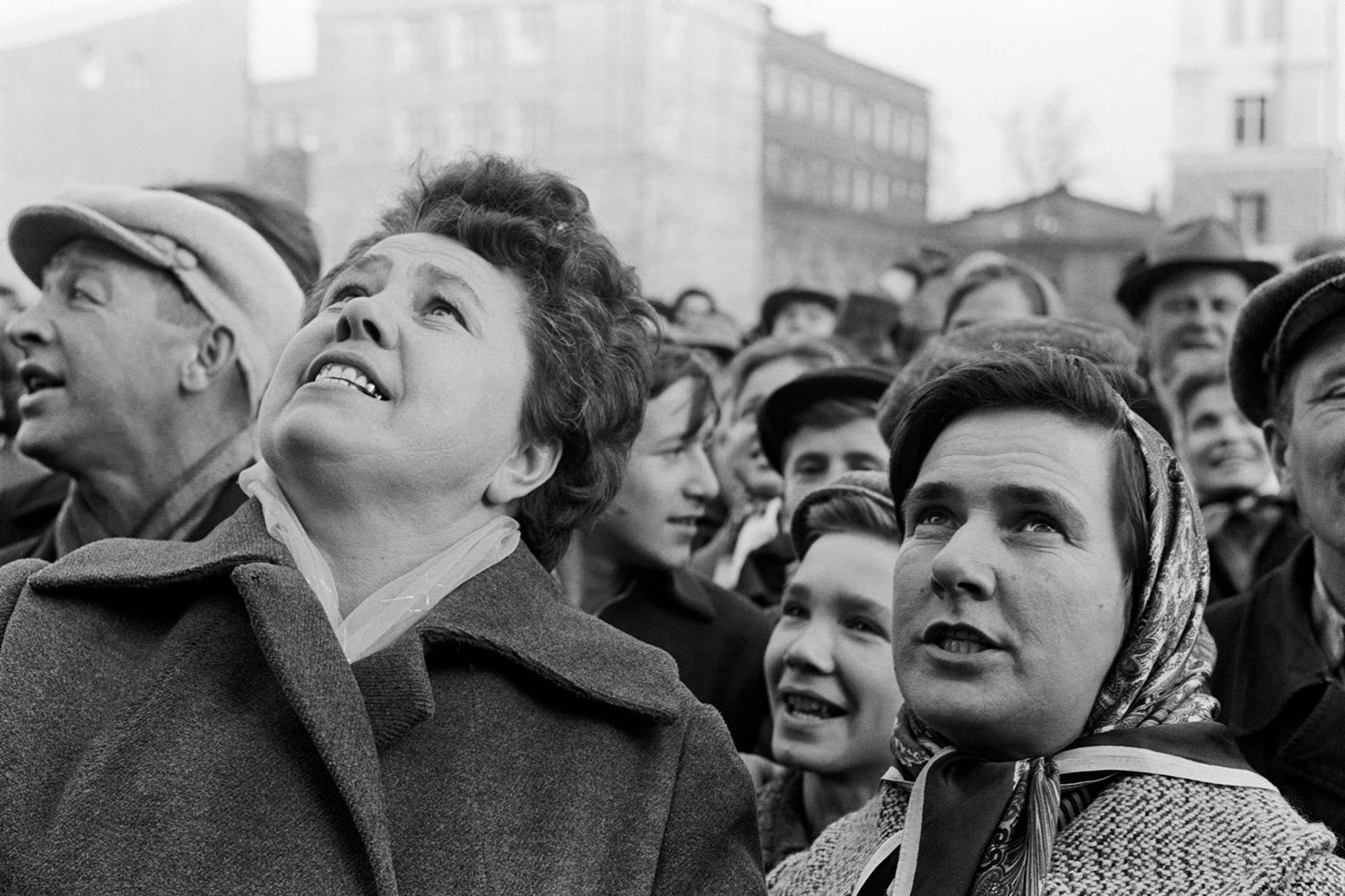 A crowd of people stand outside, some wearing hats, scarves, and overcoats, and look upward.