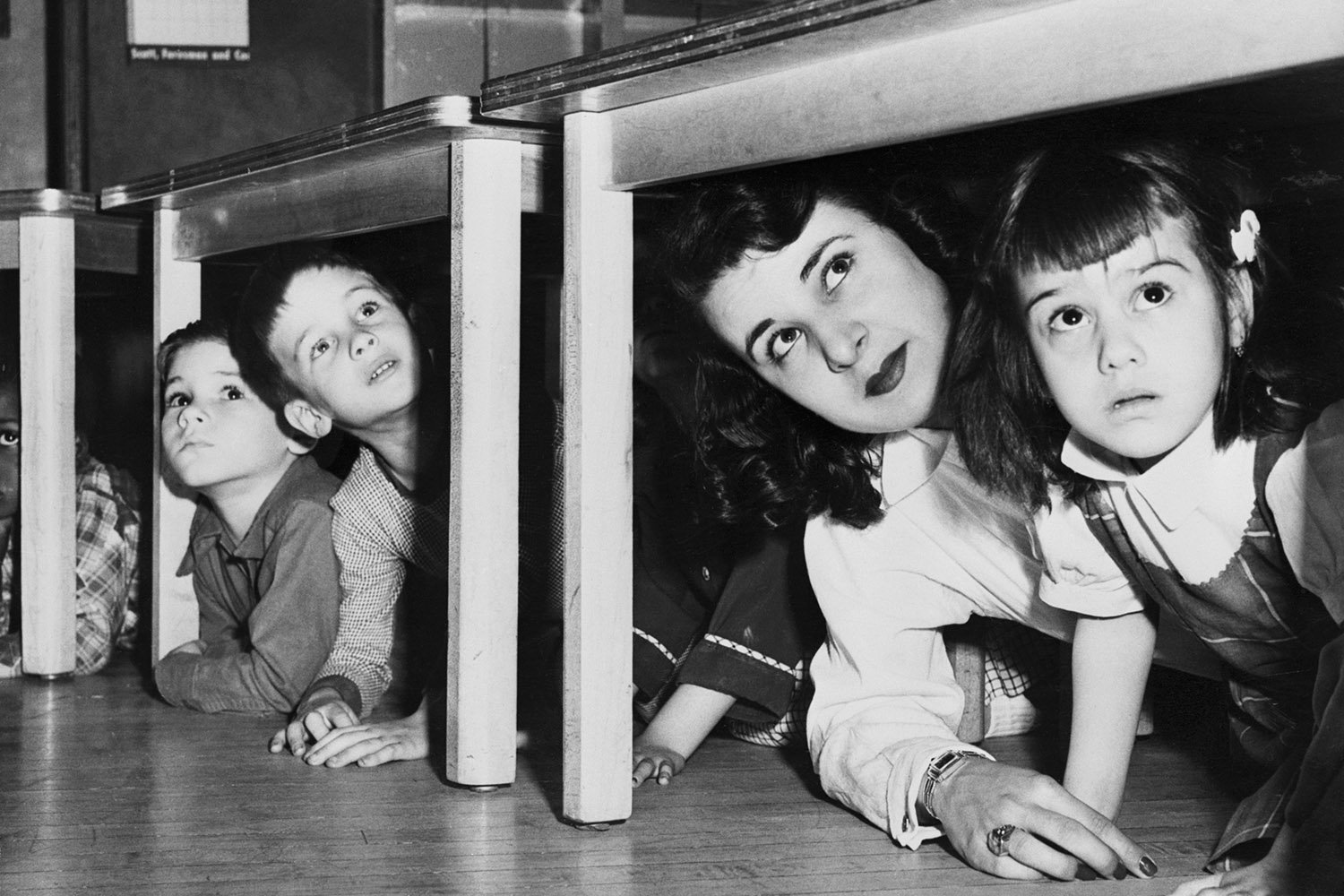 Two children peer out from under one desk while a teacher and another child peer out from another.