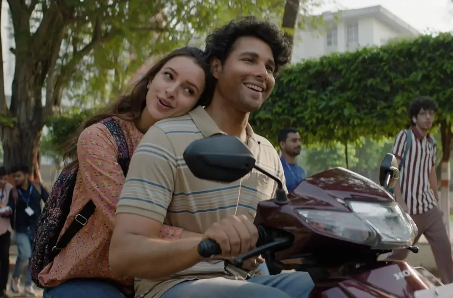 A man drives a scooter with a woman behind him, arms around his waist and head on his shoulder.
