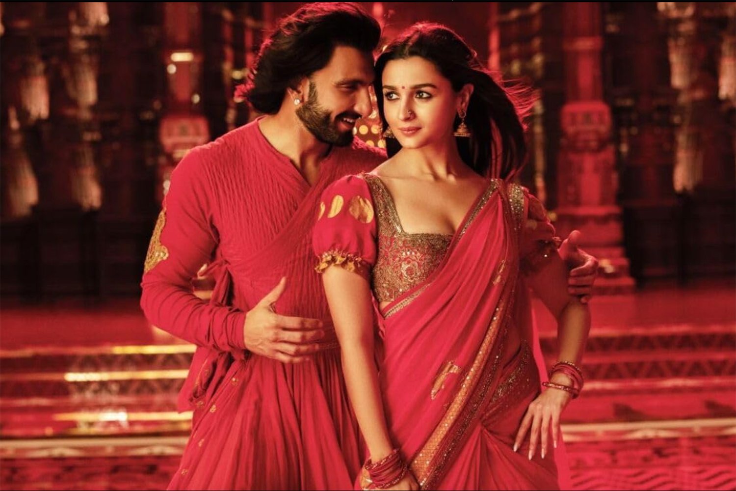 A man stands behind a woman with his hands on her shoulder and waist. They wear ornate red and gold clothing.
