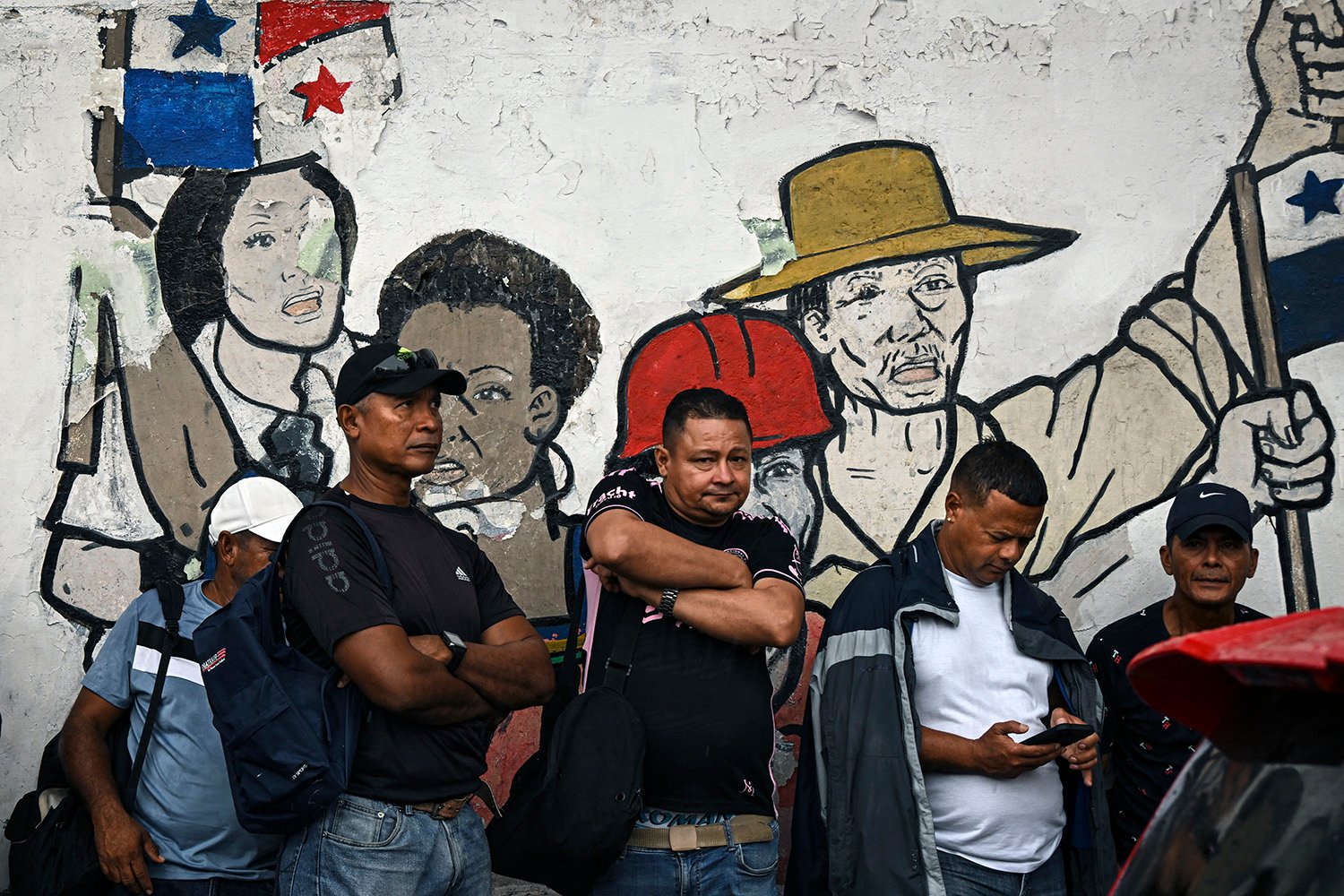 Workers stand with their arms crossed in front of a mural showing people in hats with their fists raised.