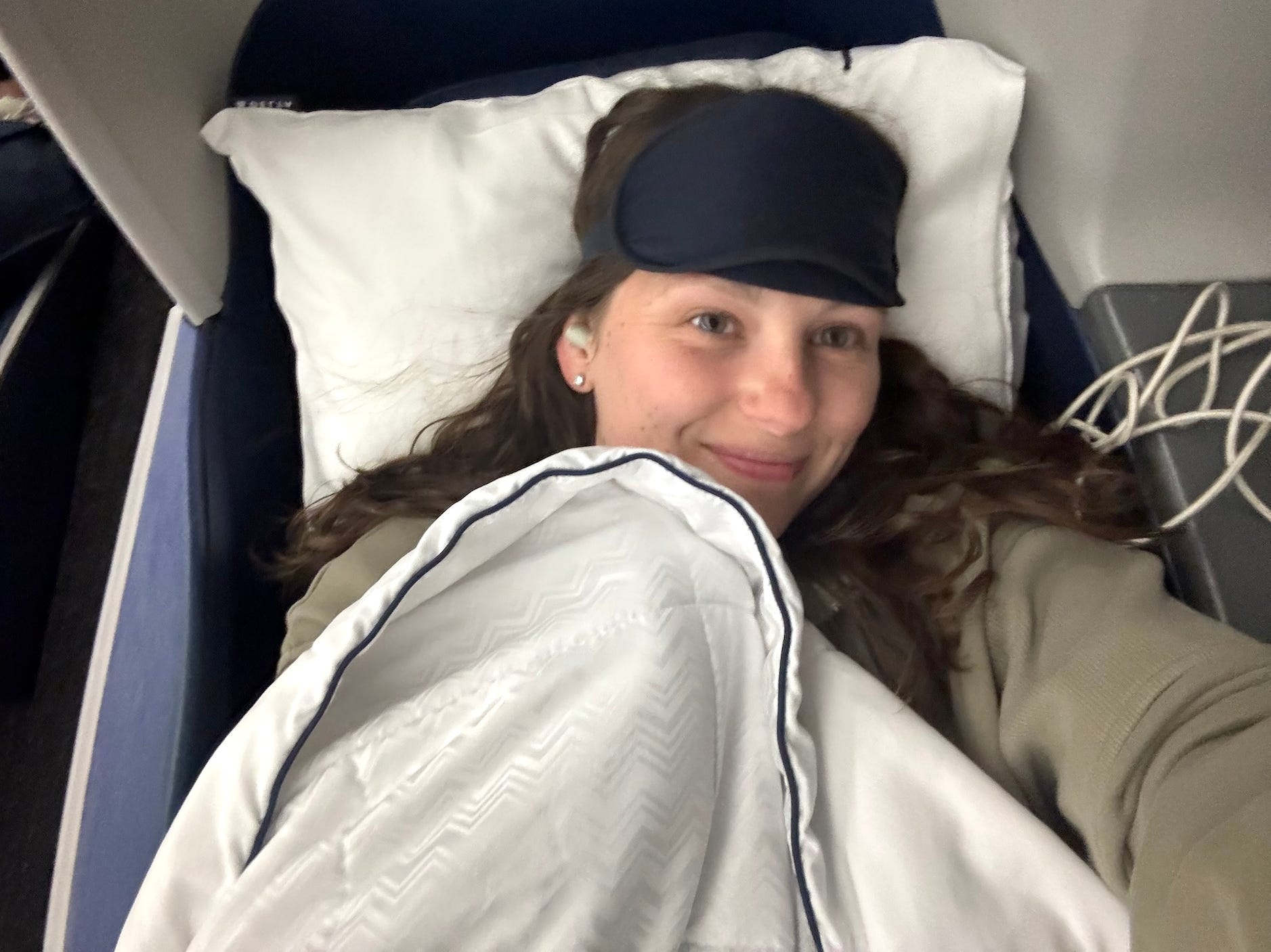 A selfie of a woman getting ready to sleep on a plane. She wears a face mask and has a pillow and blanket.