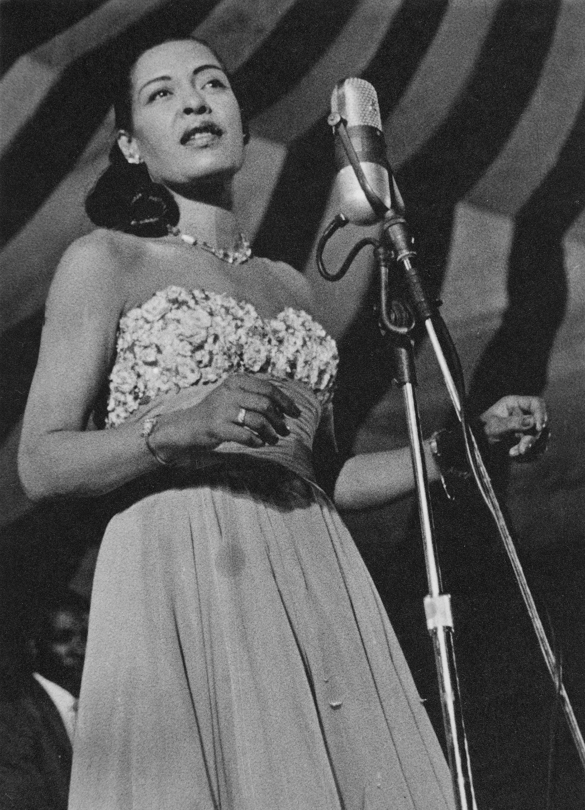 black-and-white photo of woman in strapless long cocktail dress and jeweled necklace, standing at large microphone with striped awning in background