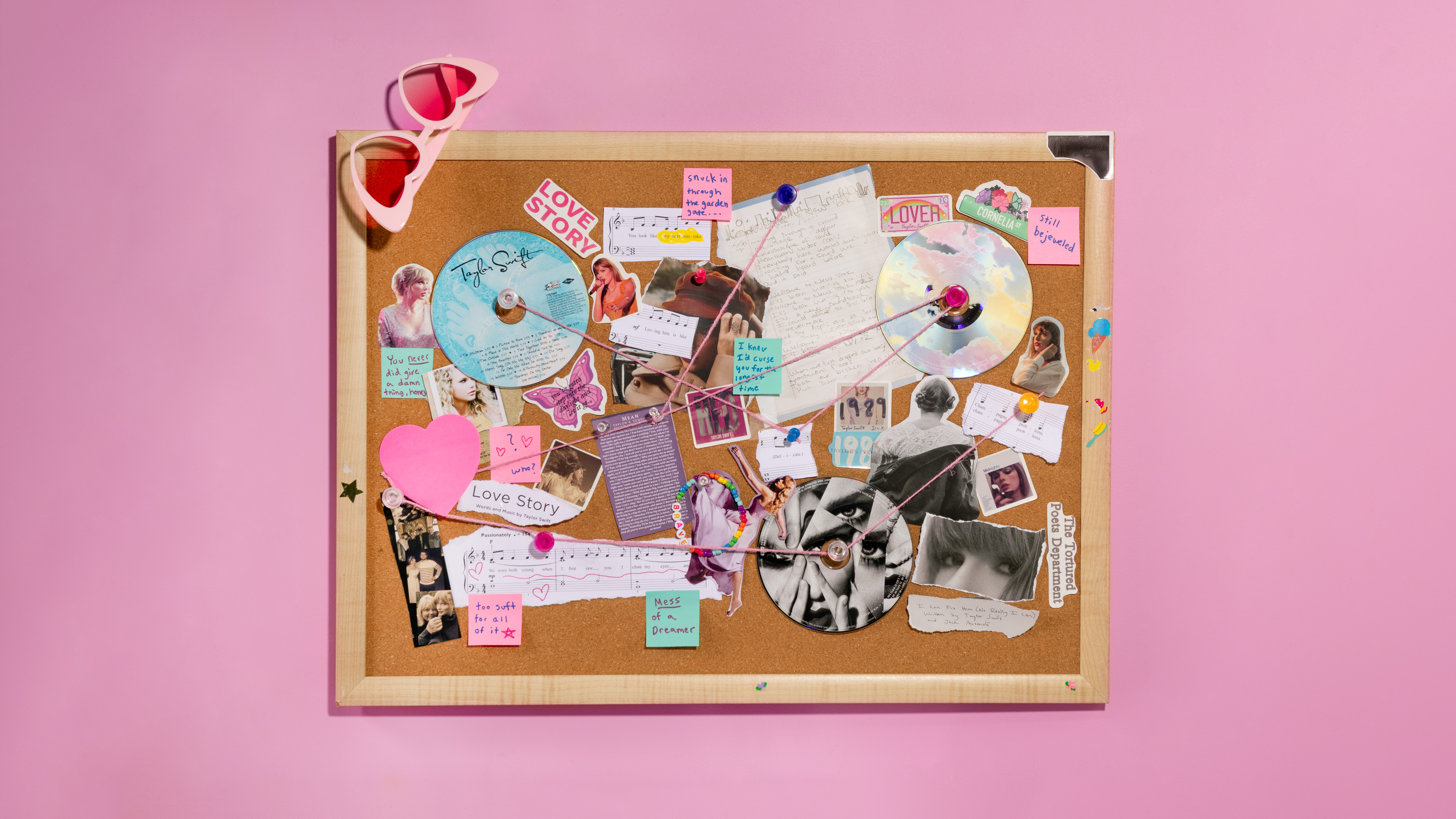 A cork bulletin board covered in Taylor Swift CDs, photos, and ephemera connected with pink string, with heart-shaped sunglasses hanging from one corner