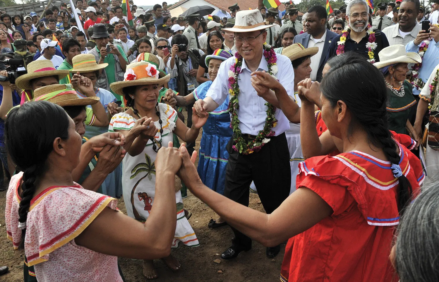 A man in a hat with flowers around his neck holds hands and dances with indigenous women in dresses in a circle amid a crowd of people.