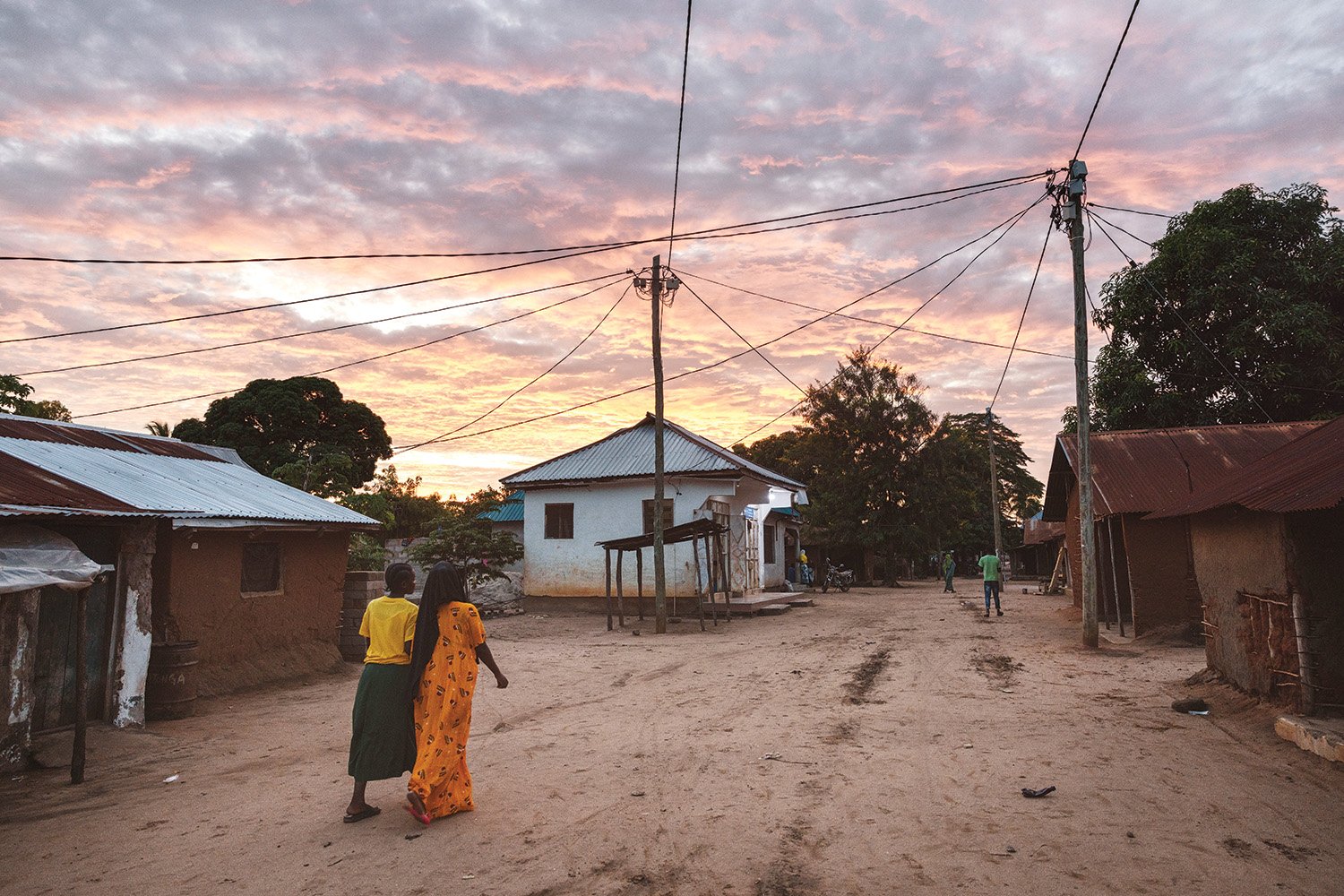 Two people walk down a dirt road under a tangle of power lines. Low buildings are seen along the road against a pink cloudy sky.