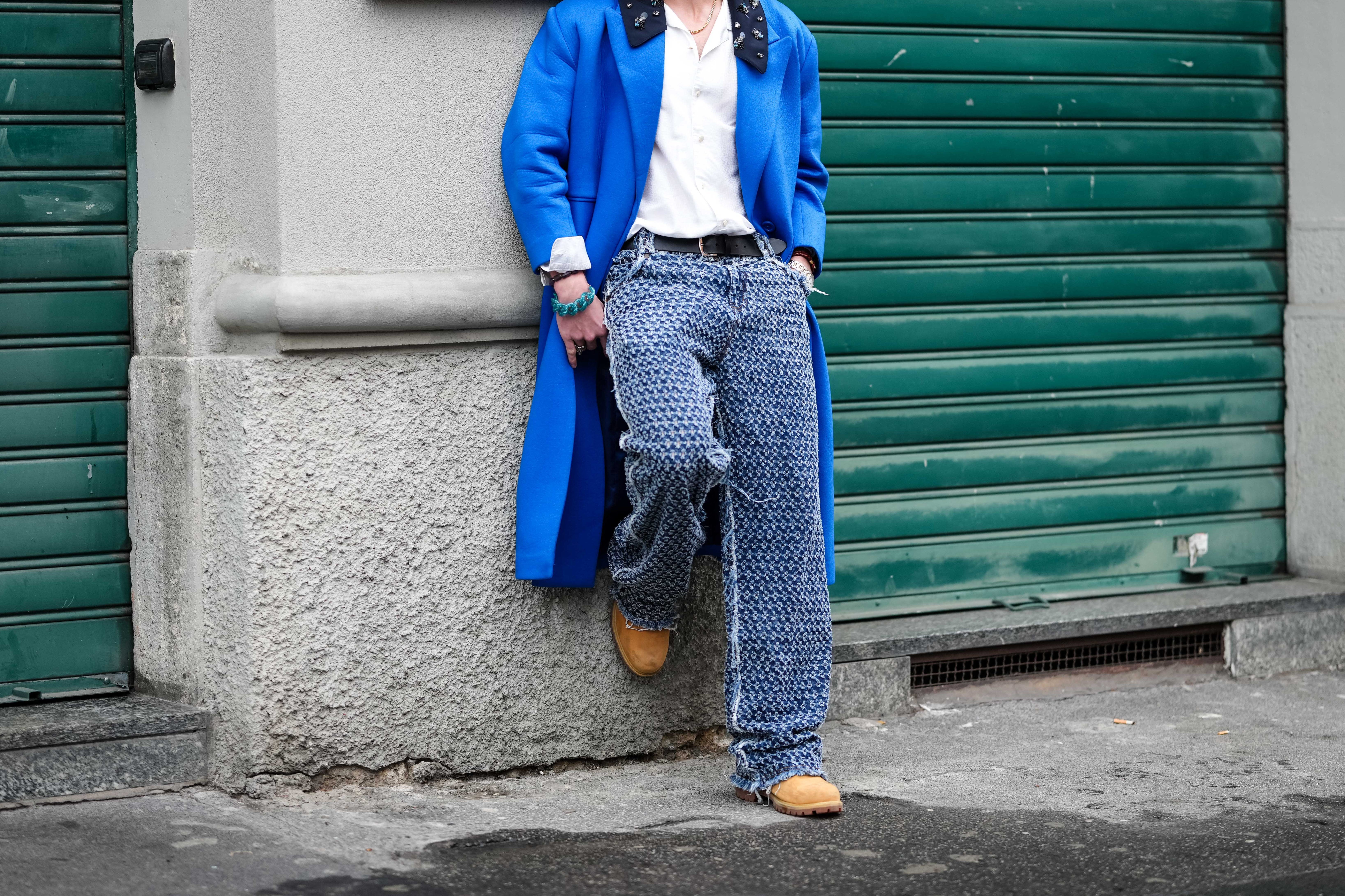 A guest wears black sunglasses, white buttoned up long sleeve shirt, bright blue coat with black collar and black detail, dark blue pattern/print loose denim jean pants, light brown boots Timberland shoes, outside Dhruv Kapoor, during the Milan Fashion week Men's Fall/Winter 2025-2026 on January 19, 2025.