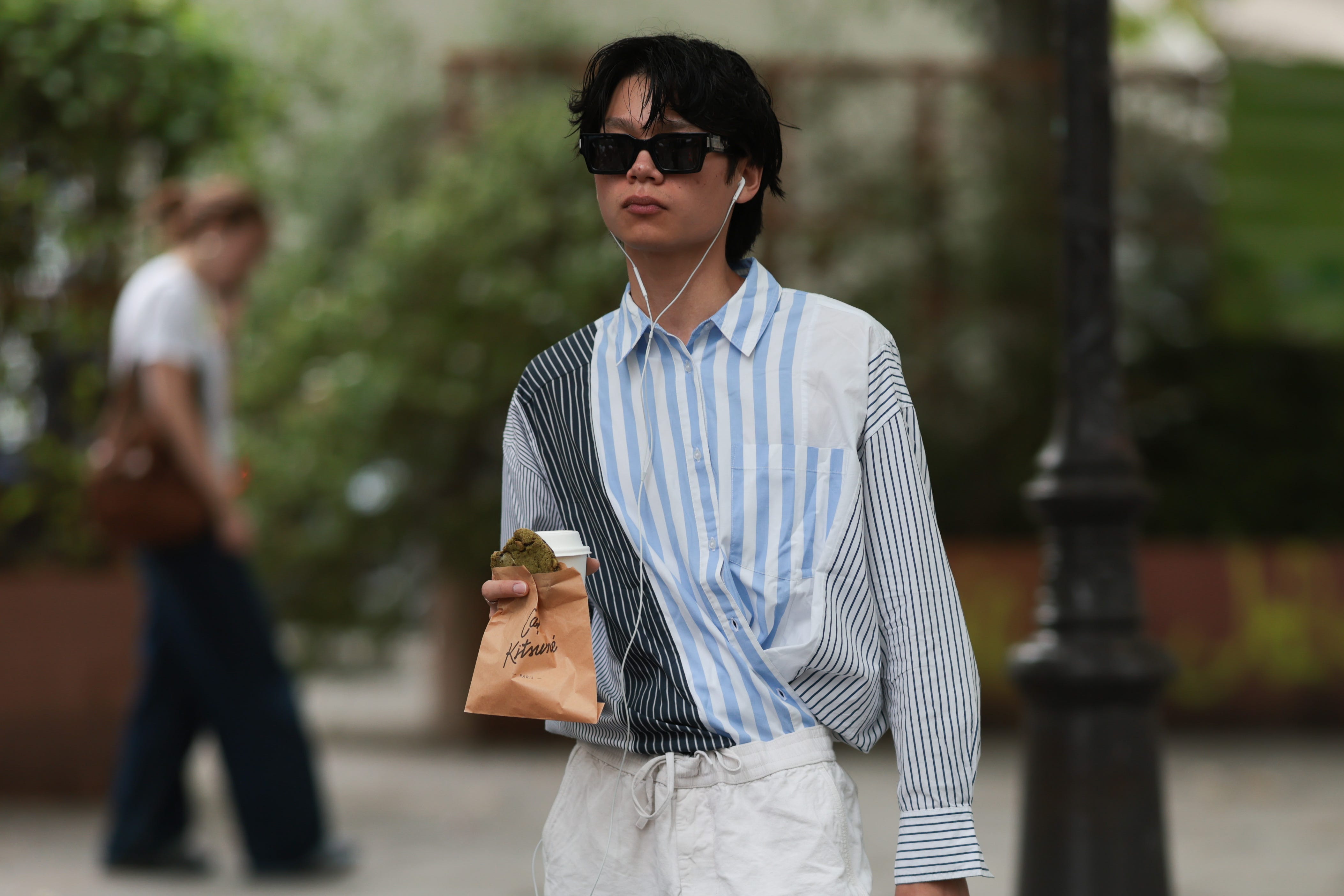 A Fashion Week guest is seen wearing rectangular black sunglasses with silver-adorned temples from Saint Laurent; a long-sleeved button-up shirt featuring irregular sections of varying striped patterns in black, white, and blue.
