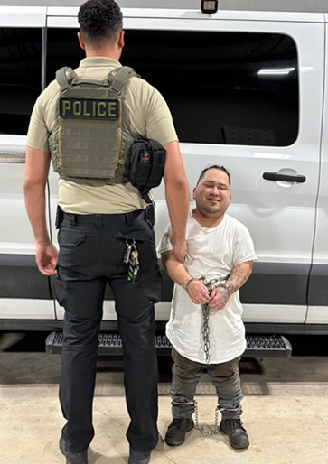 A handcuffed and chained man stands next to a police officer.