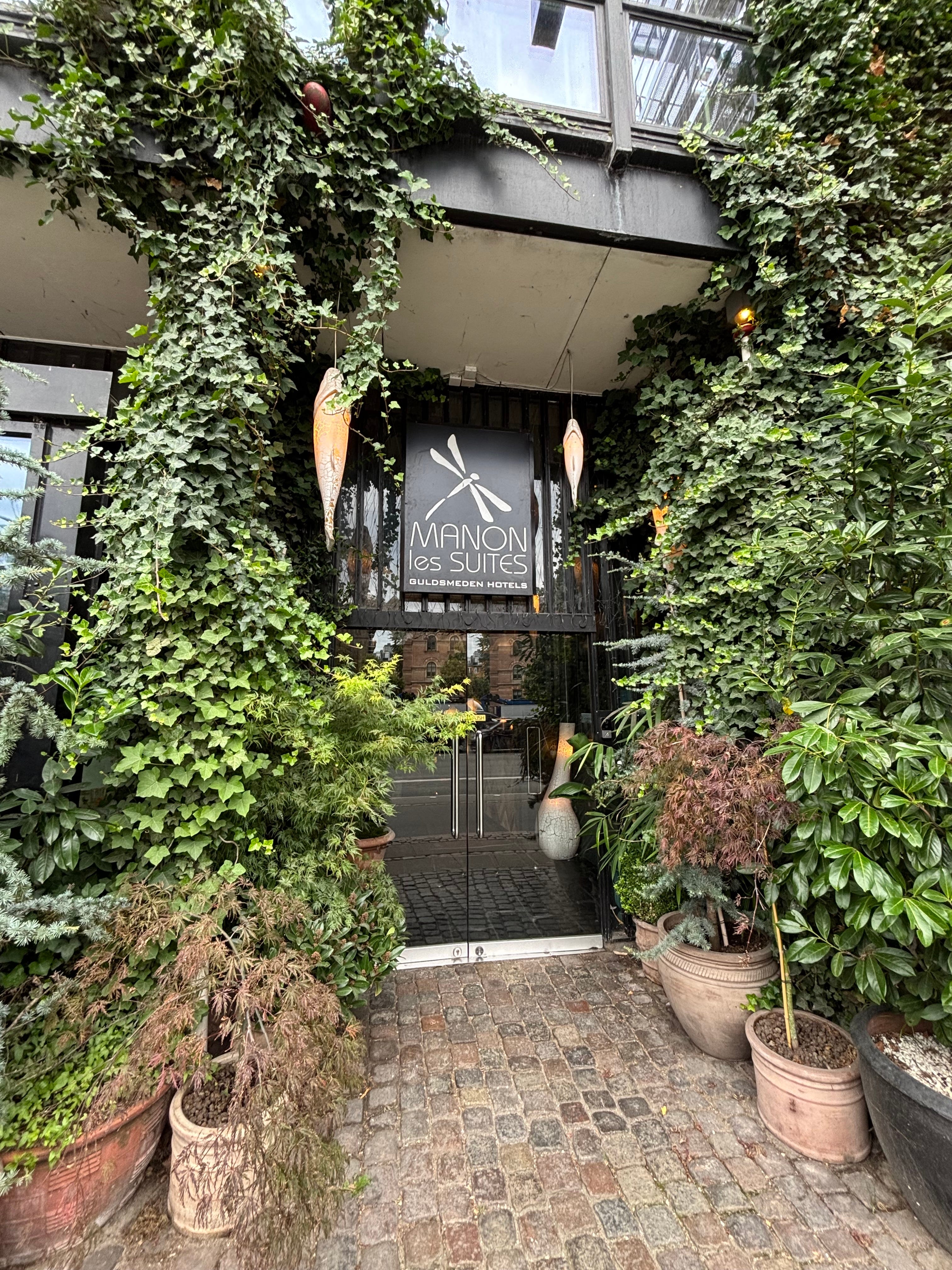 The entrance to a hotel surrounded by greenery. A sign that says "Manon Les Suites" sits above the door.