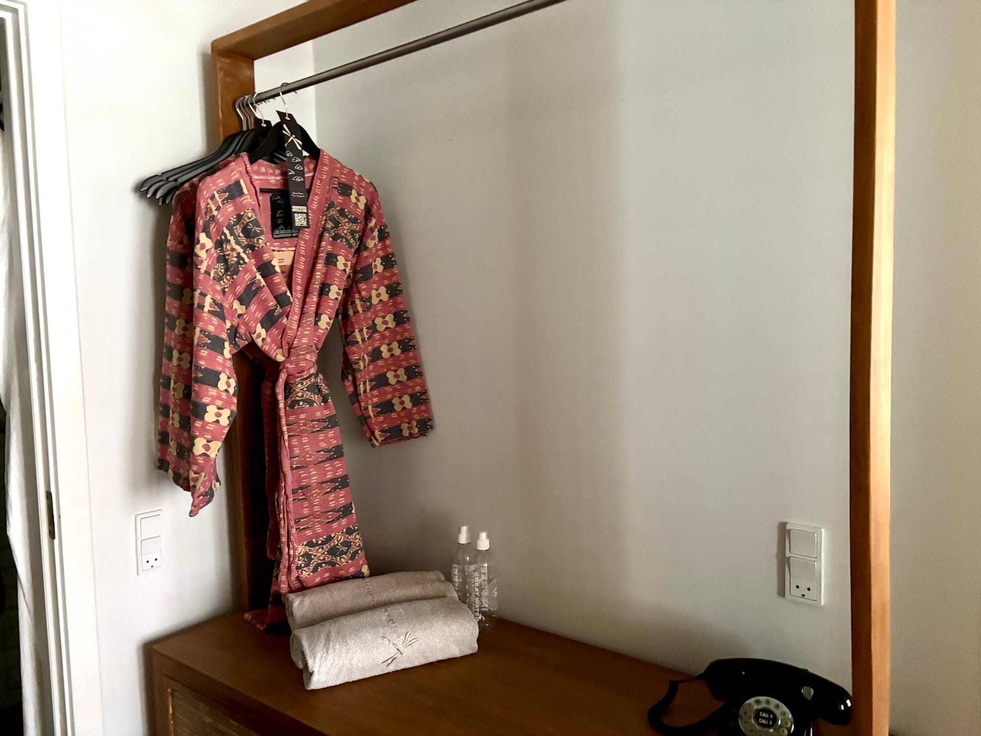 Two robes hang from a rack. Two plastic water bottles and towels rest on a dresser under them.