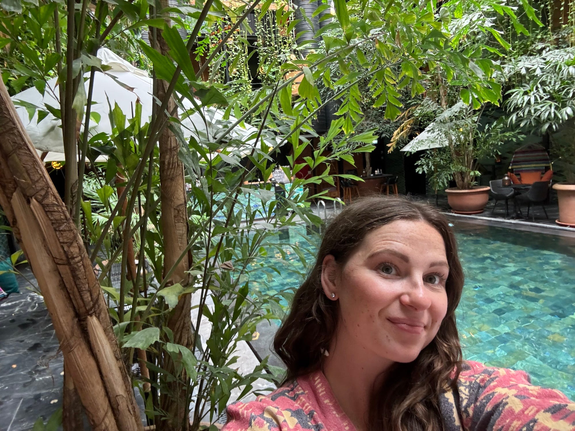A selfie of a woman in front of a pool surrounded by greenery.