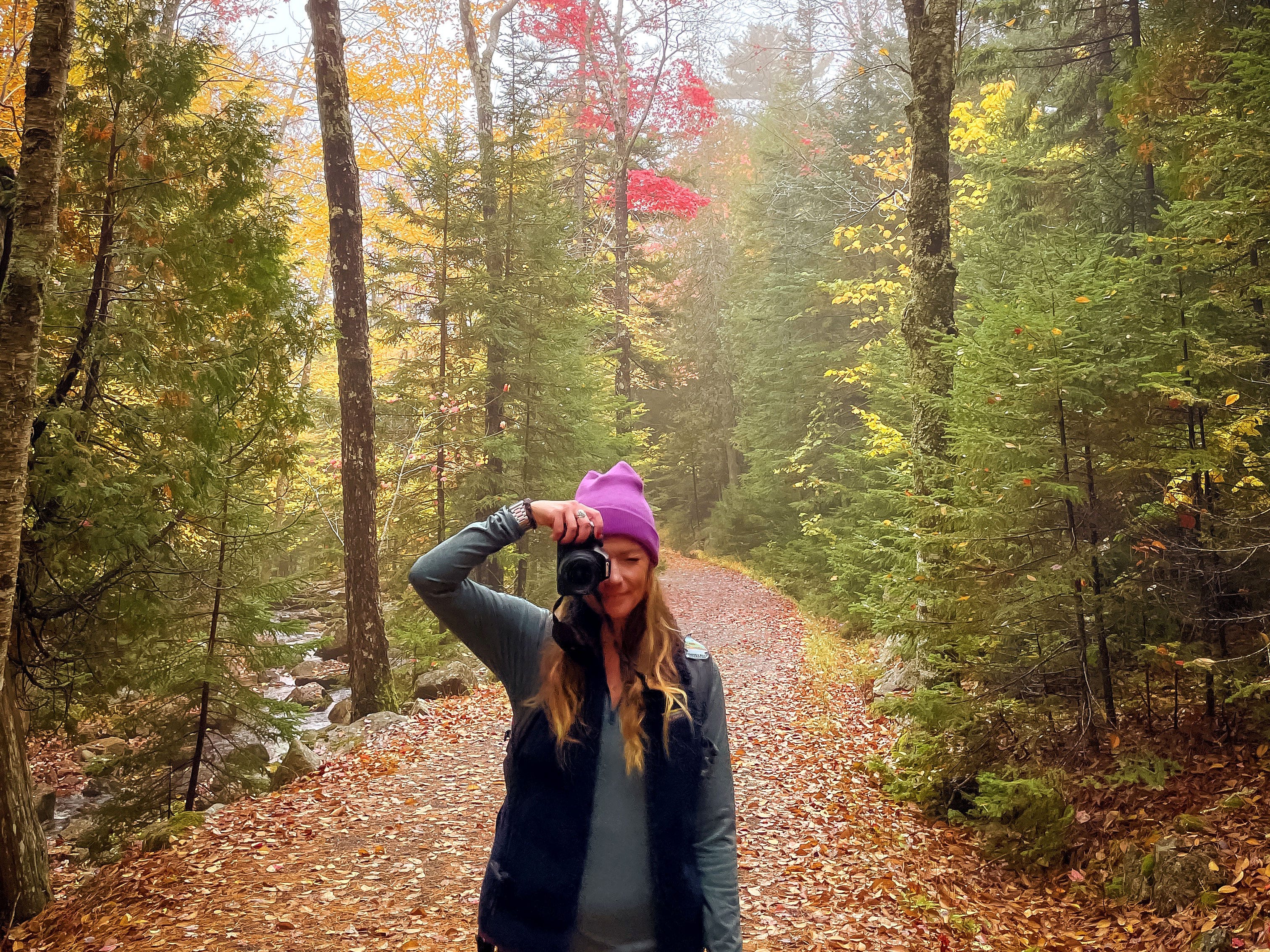 Emily holds up a camera while surrounded by colorful trees and fallen autumn leaves.