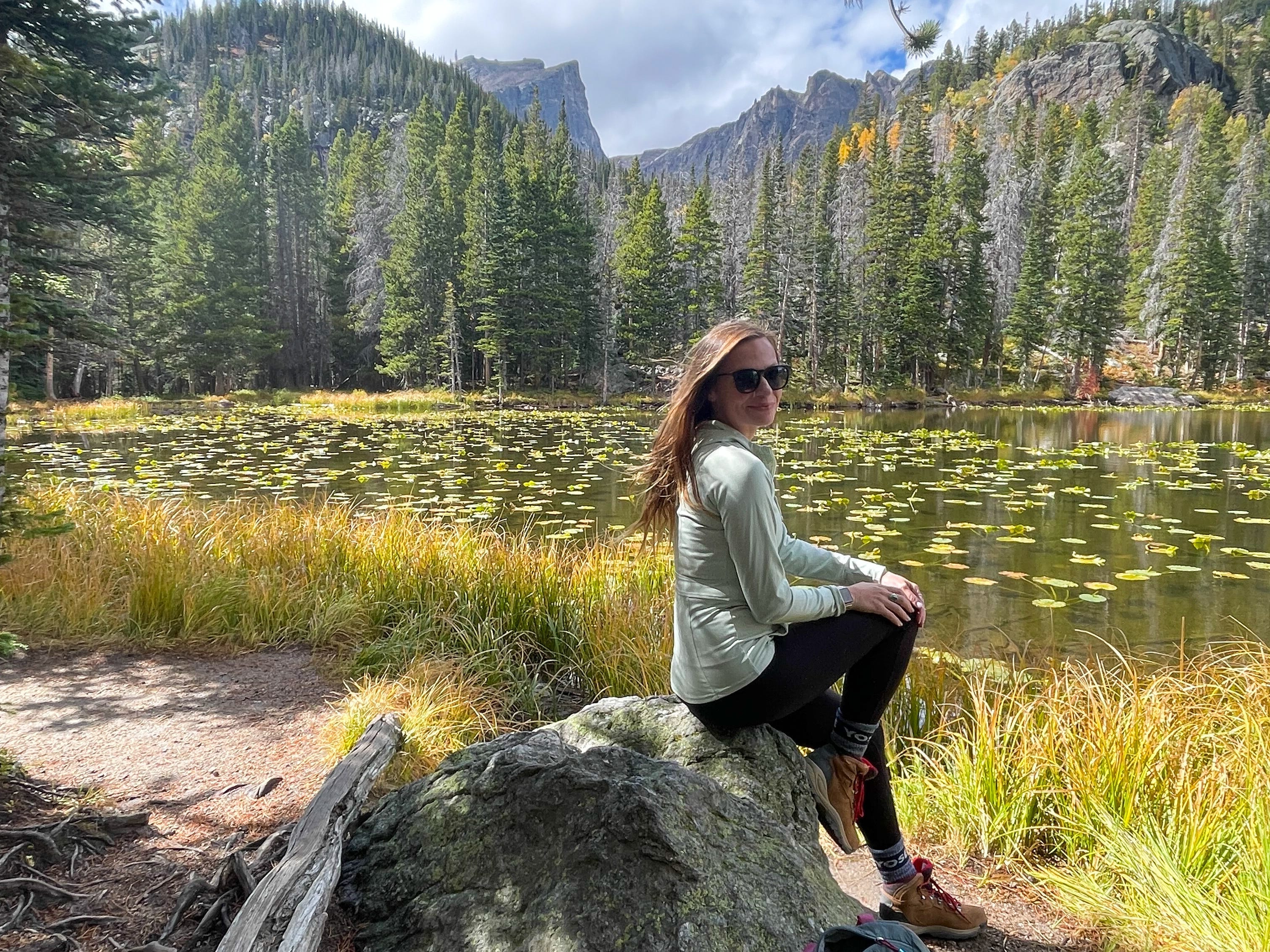 Emily sits on a rock with a lake and trees in the background in Rocky Mountain National Park.