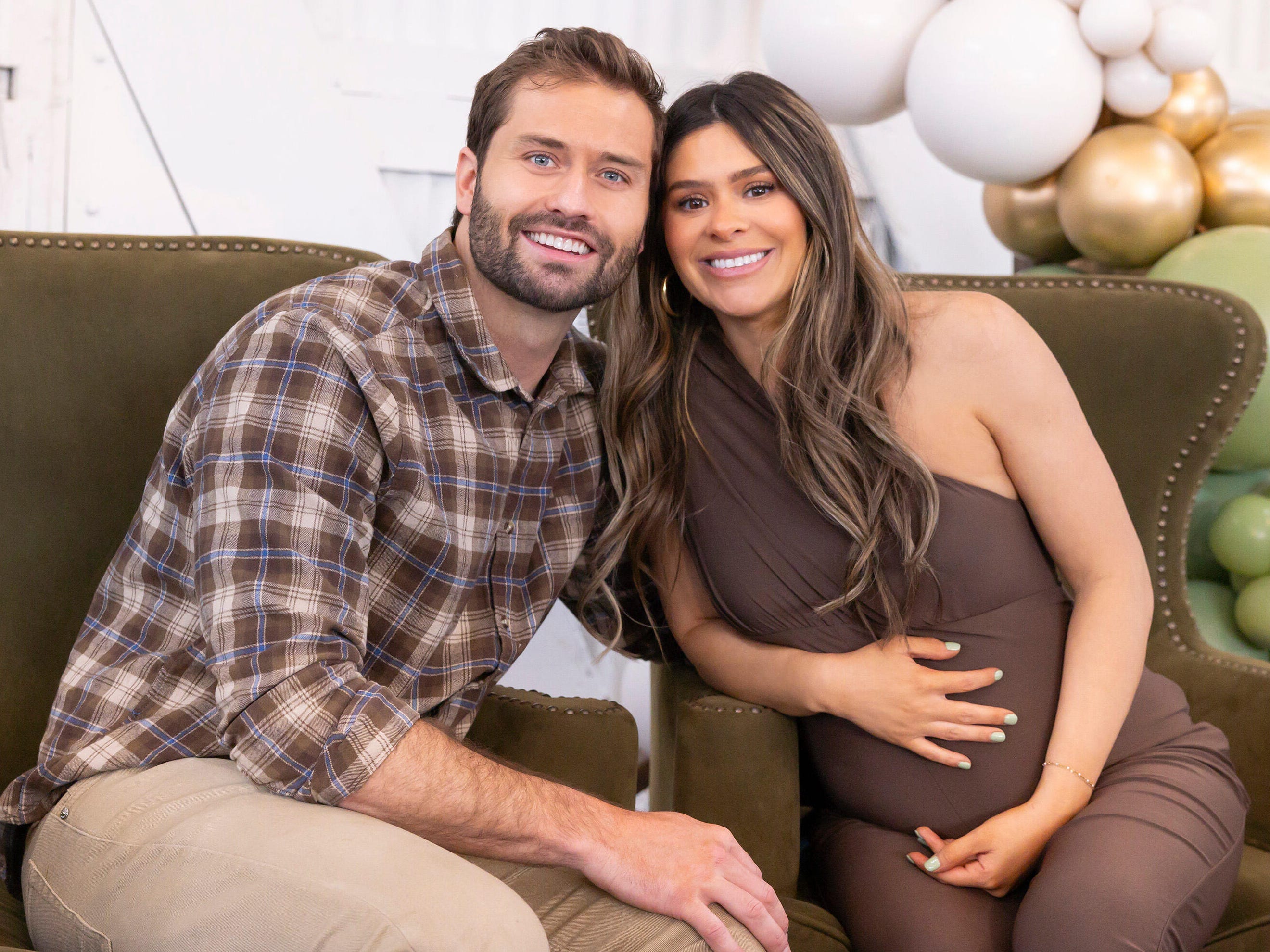 A still from "The Secret Lives of Mormon Wives" featuring Dakota Mortensen in a plaid brown shirt and brown pants hugging his partner Taylor Frankie Paul, who is in a brown dress, while they sit on two green chairs.